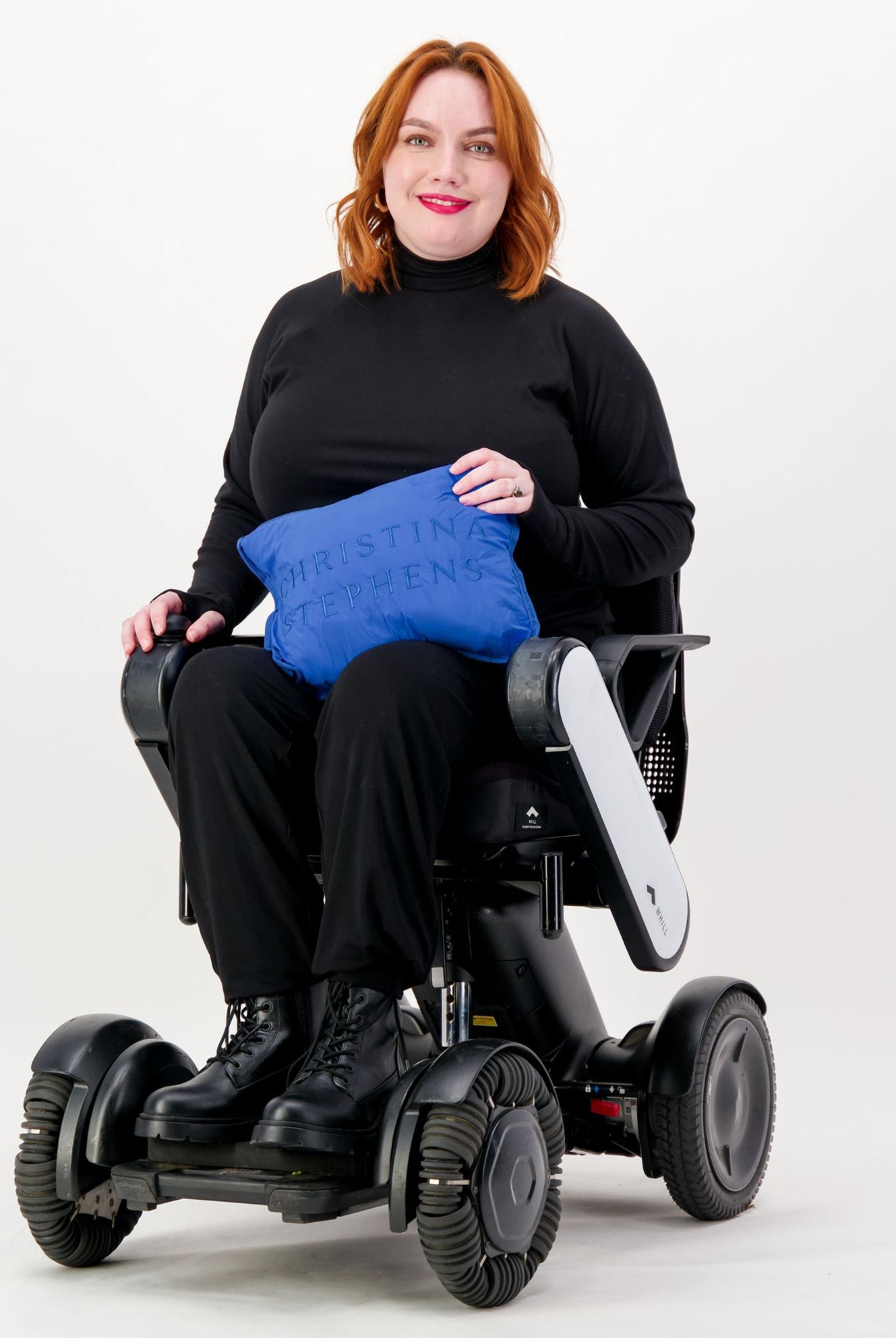 Maggie Thermal Top shown on a model seated in a modern mobility device, wearing the black turtleneck style top paired with black pants and boots, holding a bright blue cushion against a white background