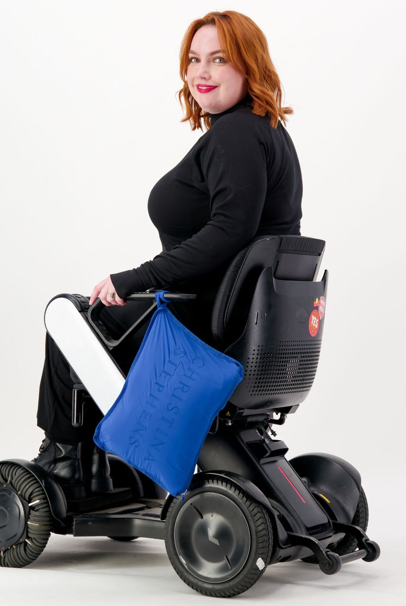 The Maggie Thermal Top shown on a model seated in a power wheelchair, featuring a classic black design with long sleeves. A bright blue accessory adds a pop of color against the sleek black garment, demonstrating its adaptable style for wheelchair users.