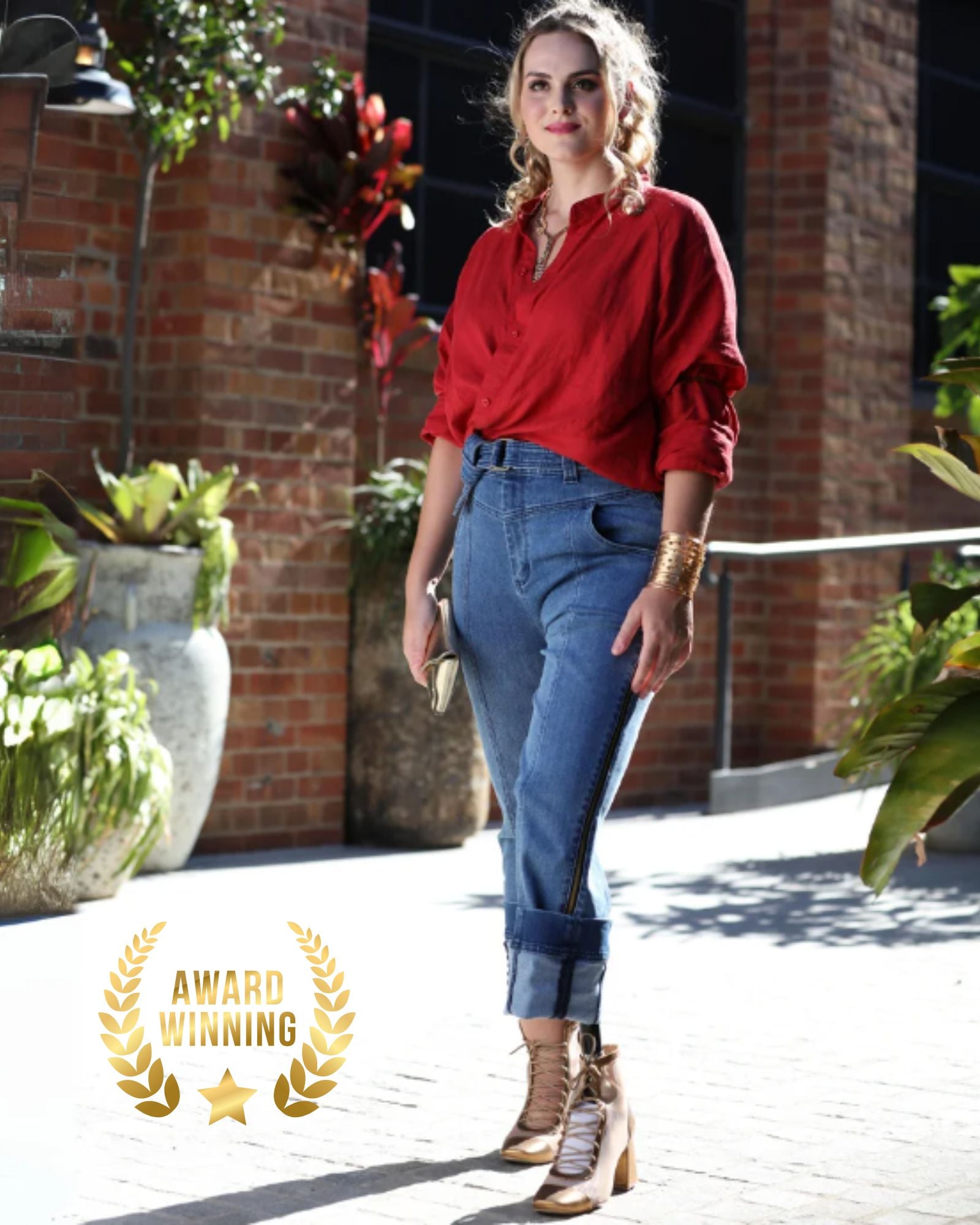 Christina Prosthetic Jeans showcased in a stylish outdoor setting, featuring a casual red blouse and award-winning adaptive denim jeans with innovative side zips, photographed against a rustic brick wall with decorative planters and natural lighting.