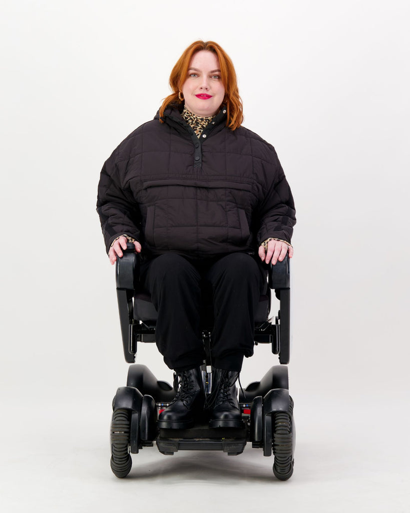 Seated female in a power chair is wearing a black puffer jacket over black pants. Christina Stephens Adaptive Clothing Australia.