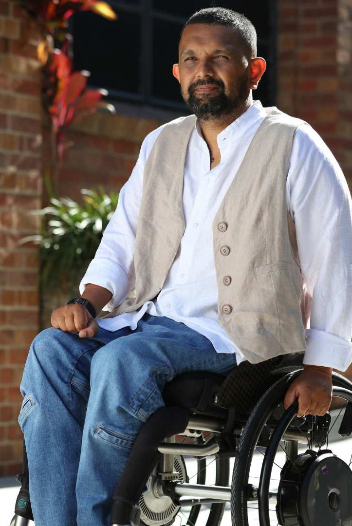 Sam Universal Linen Shirt shown outdoors against brick wall backdrop, styled with beige vest and blue jeans, demonstrating adaptive fashion design with wheelchair-friendly features in natural sunlight and garden setting.