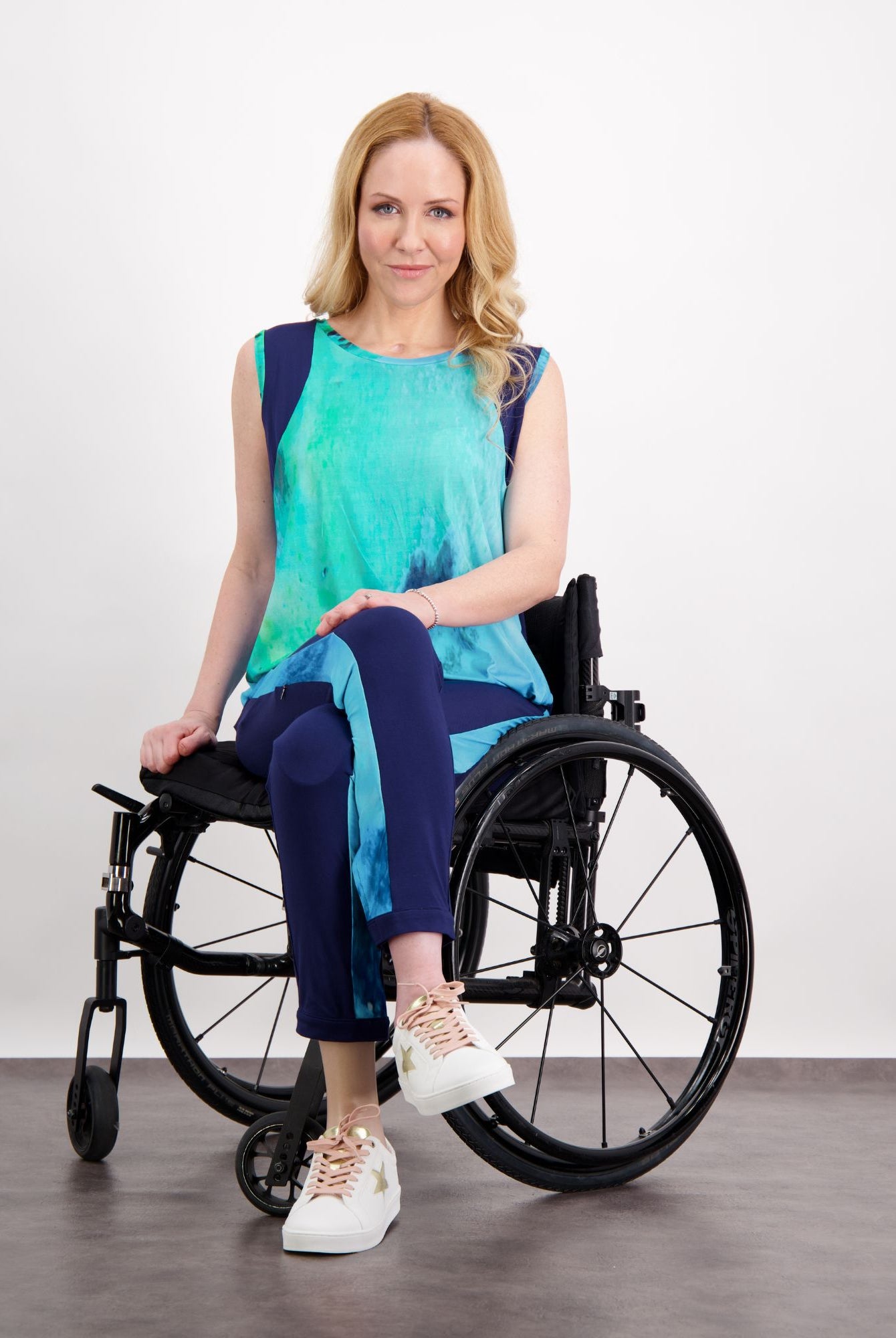 Natasha Physio Leggings shown in stylish navy blue, paired with a vibrant turquoise sleeveless top. The adaptive leggings are displayed in a seated position on a manual wheelchair, demonstrating their versatile design. White sneakers complete the modern athleisure look.