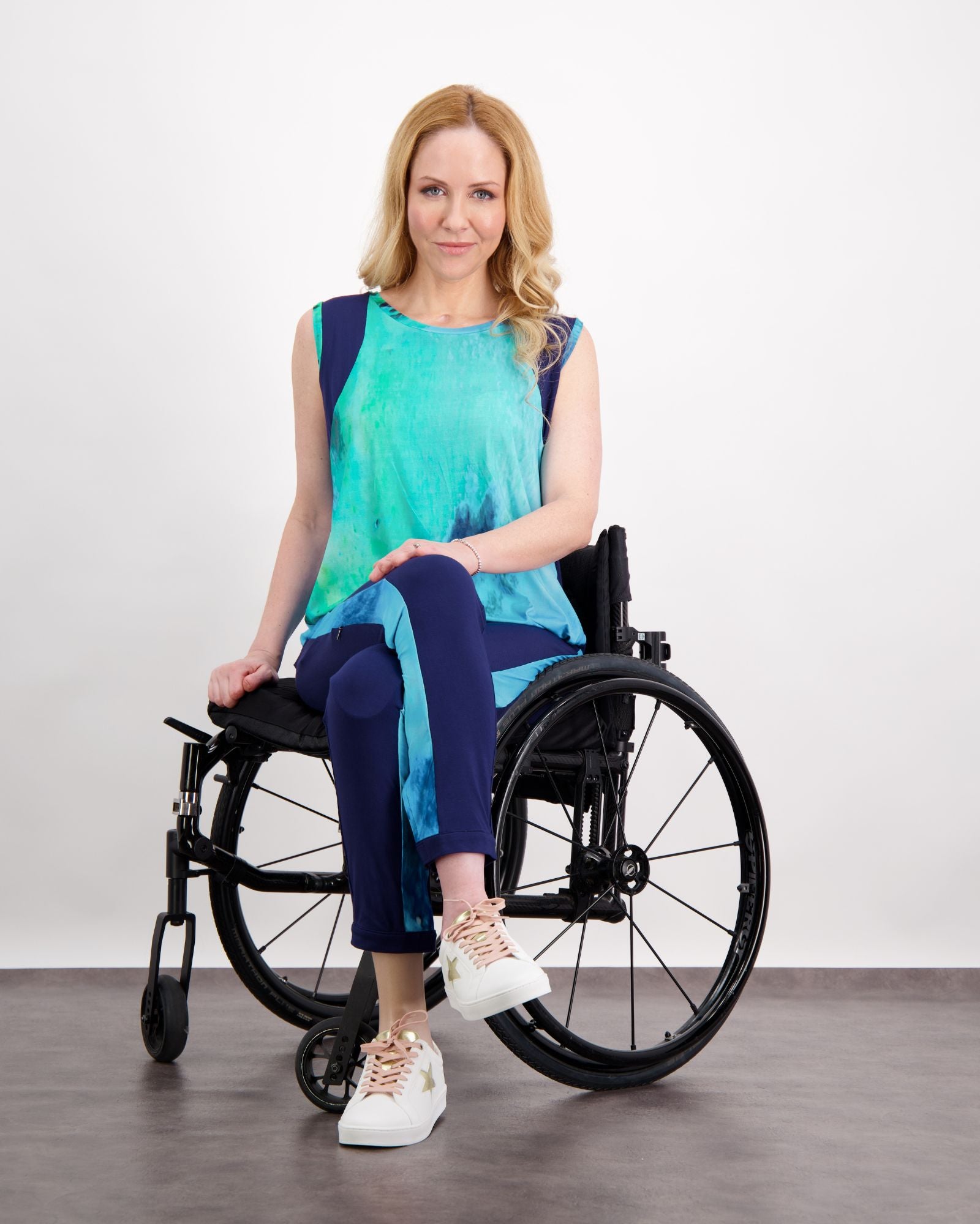 Natasha Physio Leggings shown in stylish navy blue, paired with a vibrant turquoise sleeveless top. The adaptive leggings are displayed in a seated position on a manual wheelchair, demonstrating their versatile design. White sneakers complete the modern athleisure look.