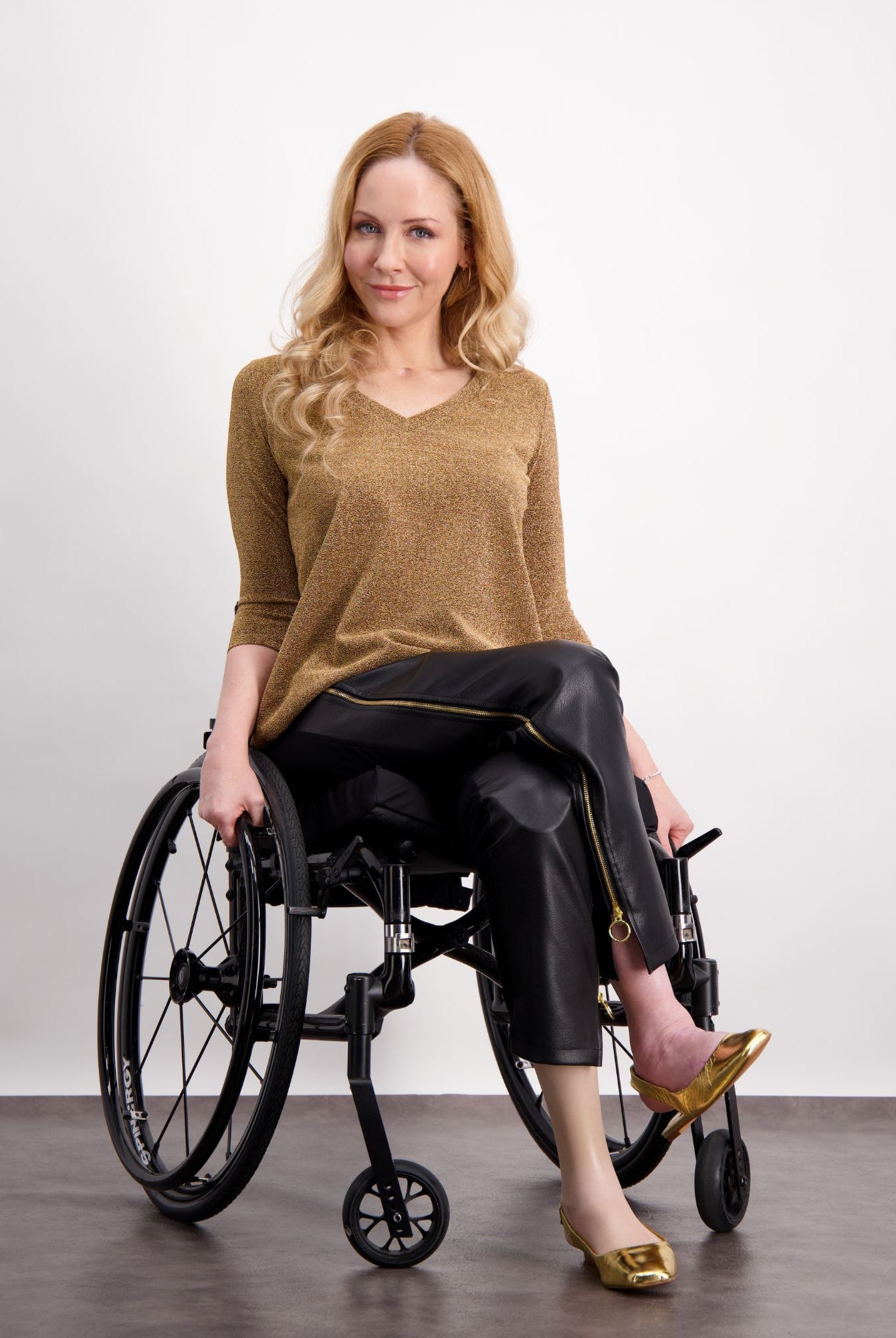Model wearing Suzie Q Seated Pants in sleek black pleather, paired with a gold V-neck sweater and metallic flats, showcasing adaptive design features while seated in a wheelchair against a light background.