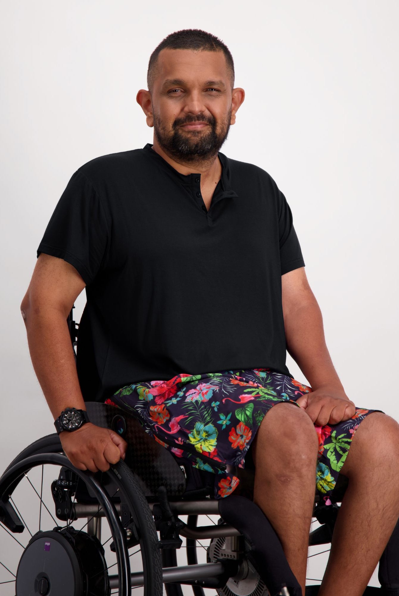 Nick A-Line T-Shirt in classic black bamboo fabric styled with colorful floral print shorts, showcasing adaptive wear comfort. Model seated in wheelchair demonstrates versatile casual wear suitable for both dressy and relaxed occasions. Product highlights sensory-friendly design and everyday comfort.