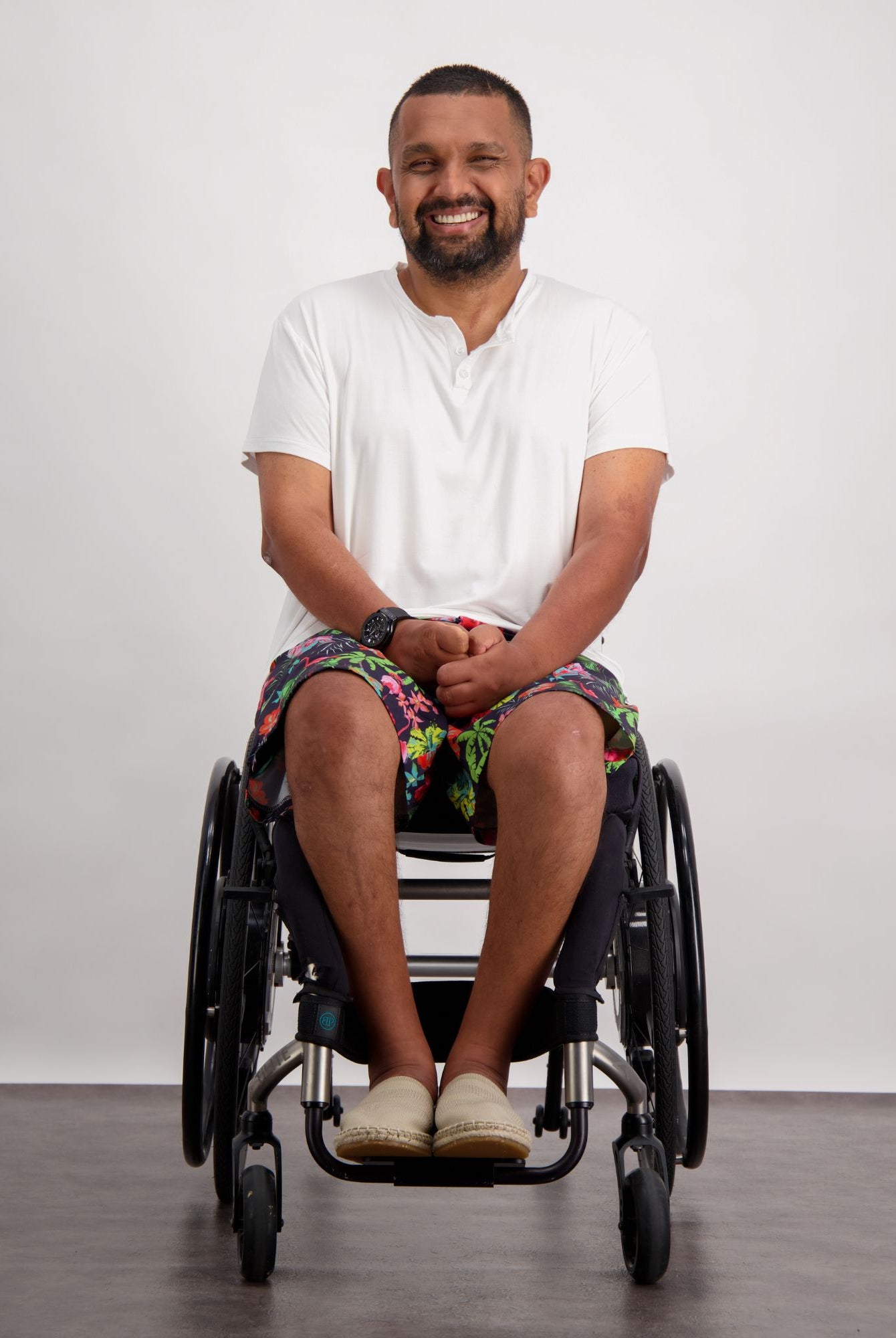 A person in a wheelchair wearing the Nick A-Line T-Shirt in white, paired with colorful patterned shorts and casual shoes. The adaptive bamboo t-shirt features a comfortable v-neck design, demonstrating its sensory-friendly fit against a neutral background.