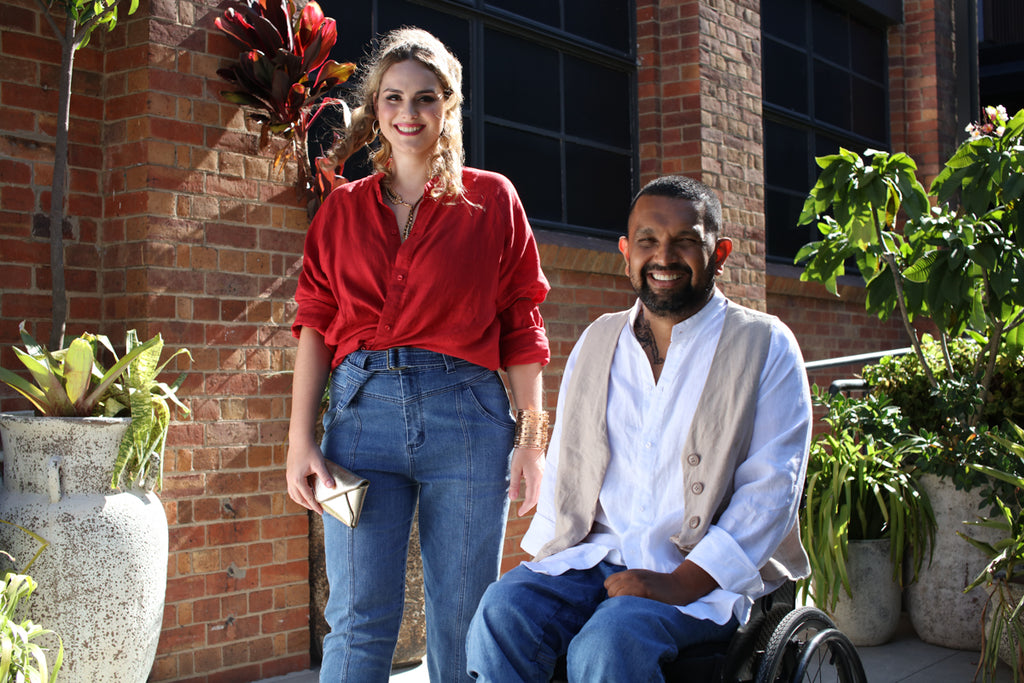 A female wearing a red shirt and blue jeans stands next to a dark skinned man in a wheelchair wearing a white shirt, beige vest and denim blue jeans. Christina Stephens Adaptive Clothing Australia. 