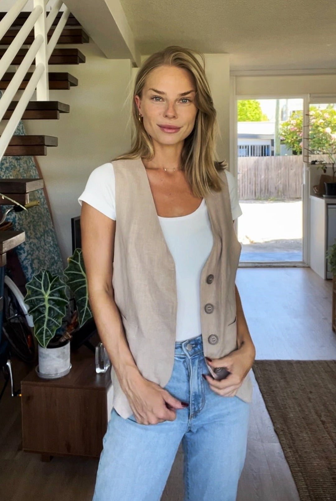 Dean Gender Neutral Waistcoat styled with white t-shirt and light blue jeans, shown in a modern home setting with floating wooden stairs and indoor plants. The beige linen waistcoat features button closures and a classic V-neck cut.