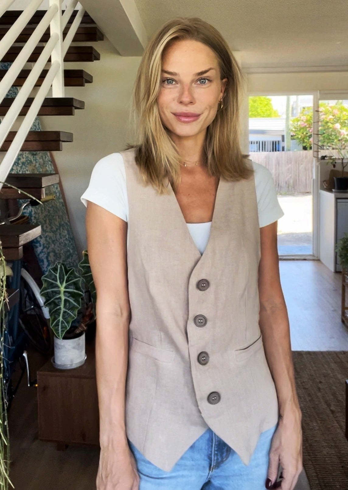 Dean Gender Neutral Waistcoat shown in beige linen fabric over white t-shirt and blue jeans, featuring asymmetrical button closure and tailored fit. Indoor setting with natural lighting and houseplants in background.