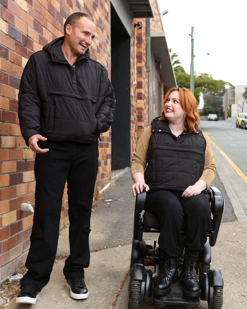 Standing male and seated female wearing midnight black puffer vest and jacket over black clothing. Christina Stephens Adaptive Clothing Australia.