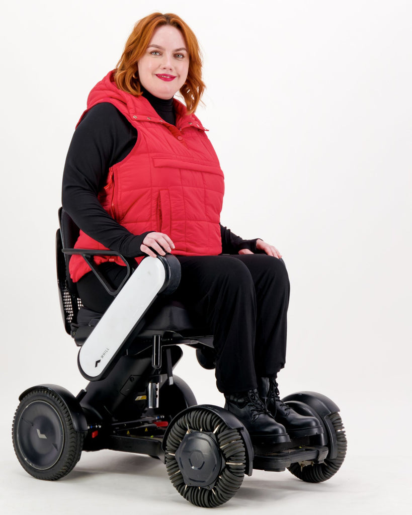 Seated female wearing a red puffer vest over black clothing while seated in a power wheelchair. Christina Stephens Adaptive Clothing Australia.