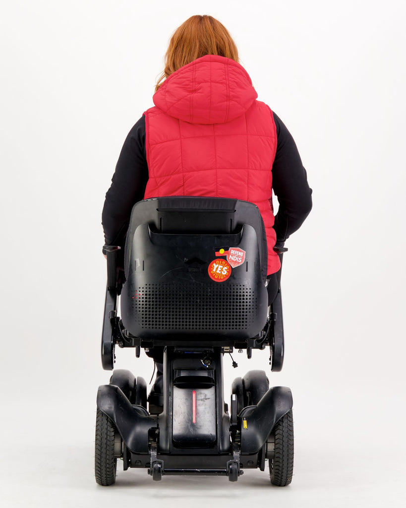 Seated female wearing a red puffer vest over black clothing while seated in a power wheelchair. Christina Stephens Adaptive Clothing Australia.