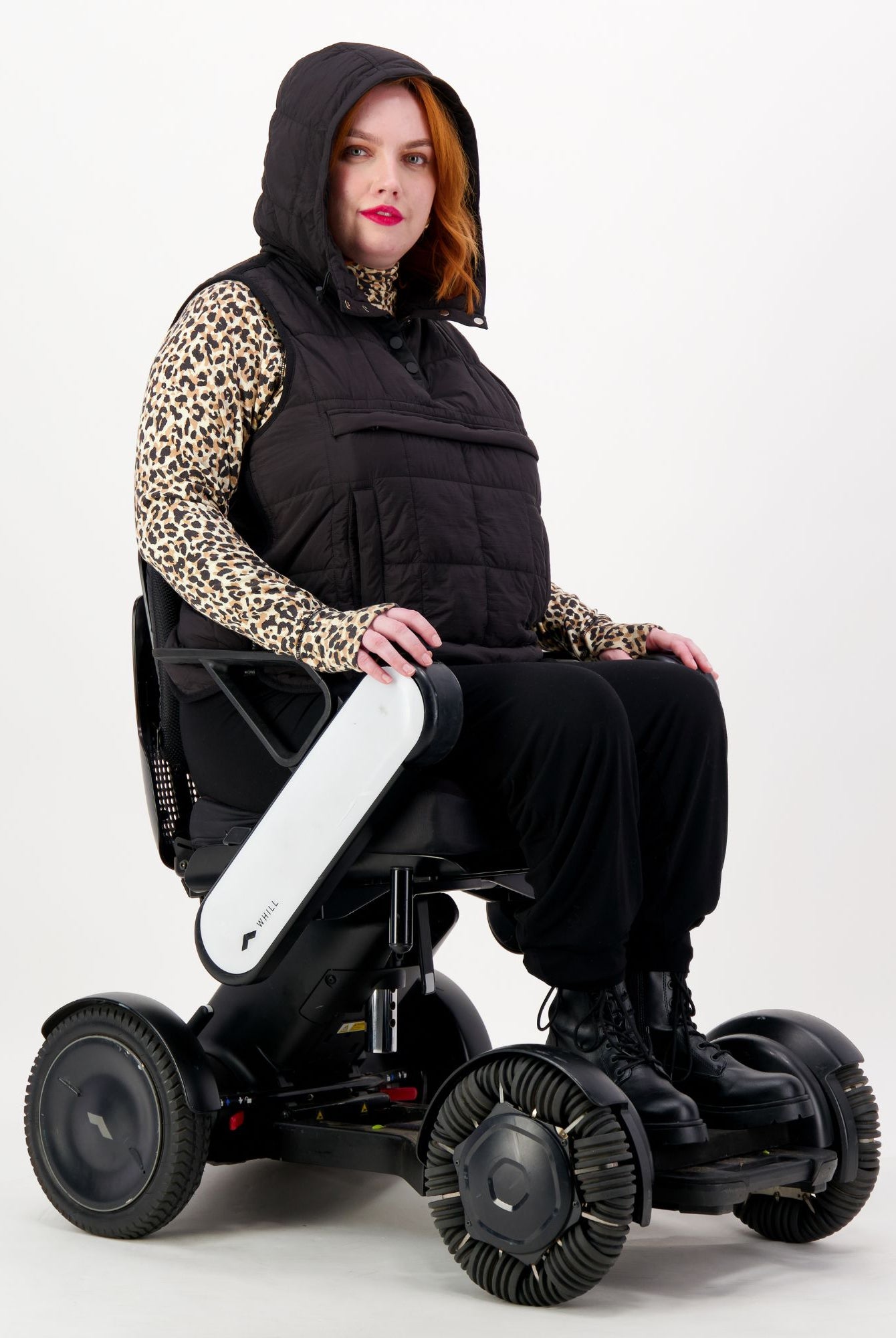 Matt Gender Neutral Puffer Vest shown on a wheelchair user wearing a leopard print top. Black hooded vest features adaptive design for wheelchair comfort. Model seated in a modern white electric wheelchair against white background.