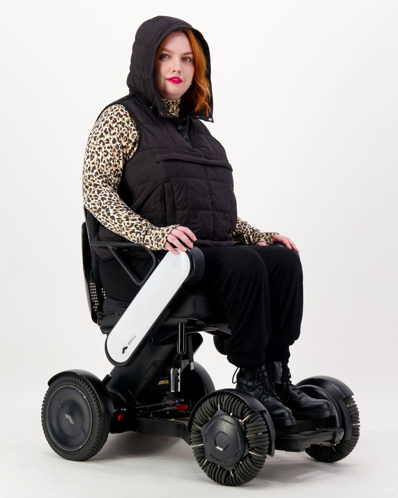 Seated female wearing a black puffer vest over black clothing while seated in a power wheelchair. Christina Stephens Adaptive Clothing Australia.