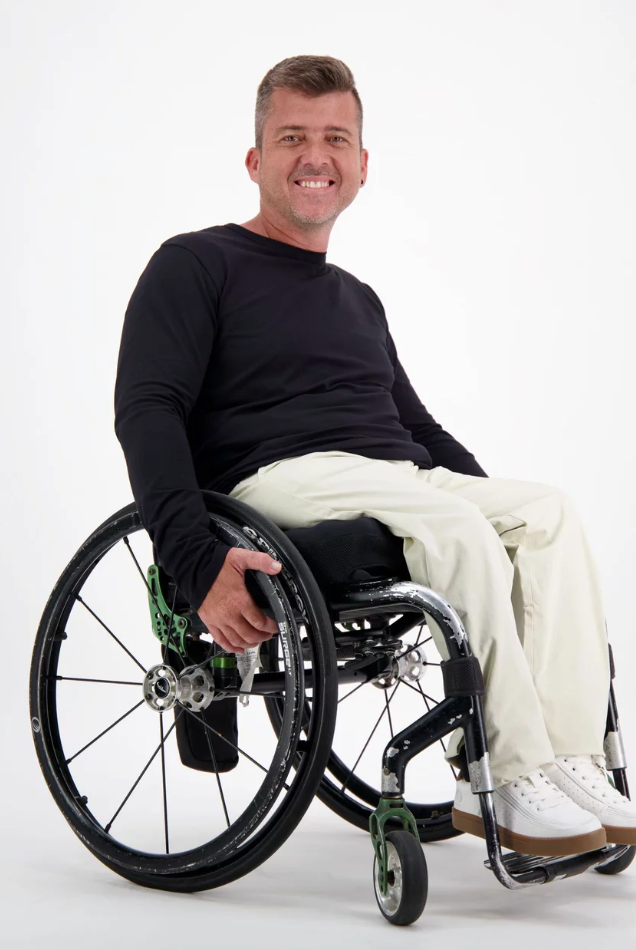Mens Long Sleeve Leaf Back T-Shirt shown on model in wheelchair against white background, featuring classic black long sleeve design, paired with white pants and sneakers. Adaptive clothing demonstrates comfortable fit and easy accessibility.