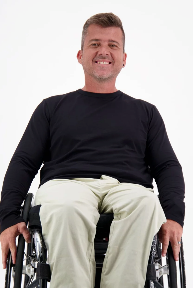 Mens Long Sleeve Leaf Back T-Shirt shown on a person seated in a wheelchair, wearing khaki pants against a white background. The black t-shirt features a classic crew neck design with long sleeves, demonstrating comfortable casual wear that's accessible and stylish.