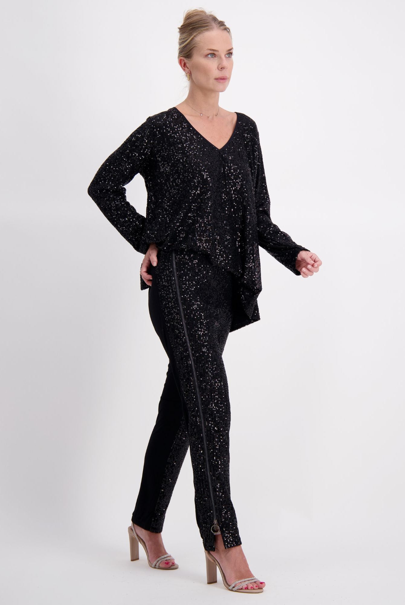 Suzie Q Seated Pants Sequins shown in elegant black sequin fabric. Model wearing matching v-neck tunic top with long sleeves, creating a stunning evening ensemble. Adaptive pants feature seamless design and accommodating fit, styled with nude heels against white background.