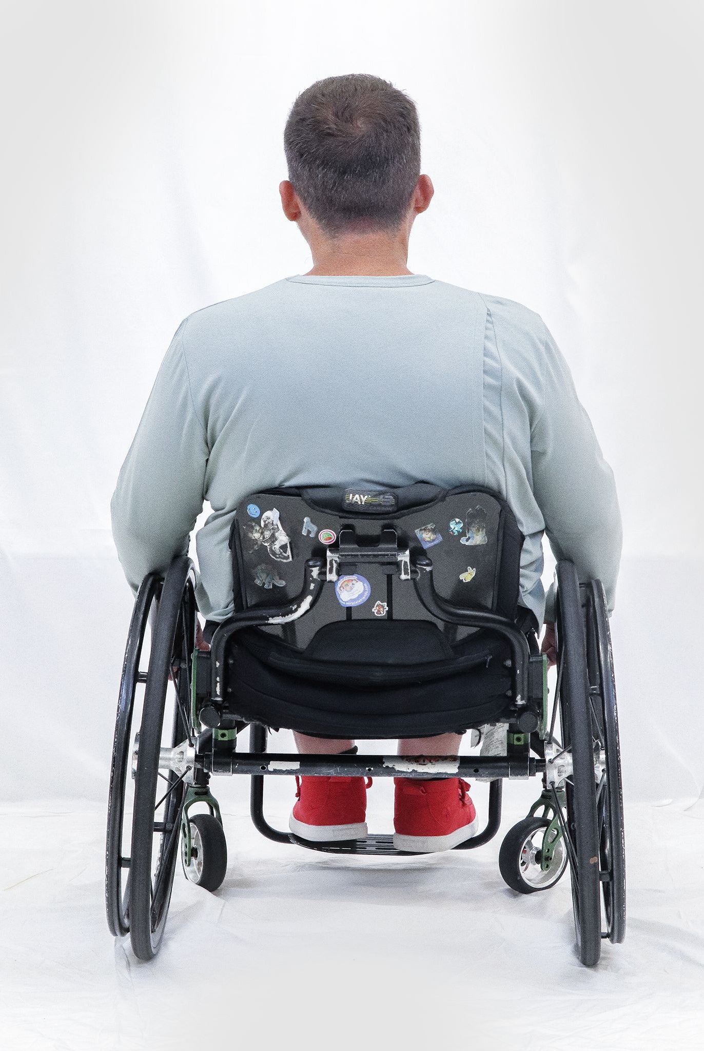 Back view of Men's Long Sleeve Leaf Back T-Shirt shown on a person in a wheelchair, wearing a light grey shirt with red shoes, demonstrating the comfortable fit and casual style against a white background.