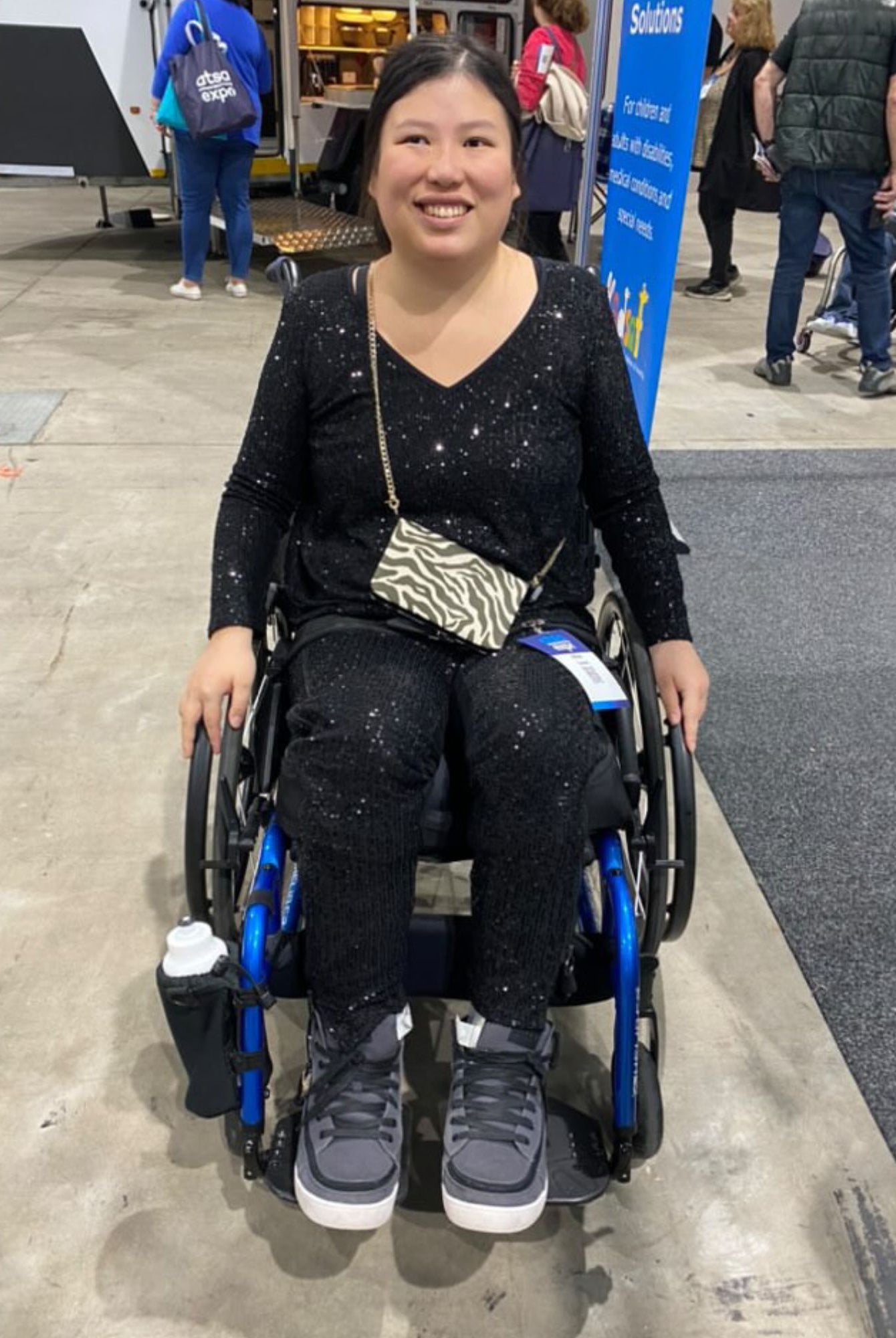 Suzie Q Seated Pants Sequins shown on a wheelchair user at an event, featuring a sparkly black sequined fabric. The adaptive pants are paired with a zebra-print crossbody bag and grey sneakers, demonstrating the stylish yet functional design perfect for wheelchair users.