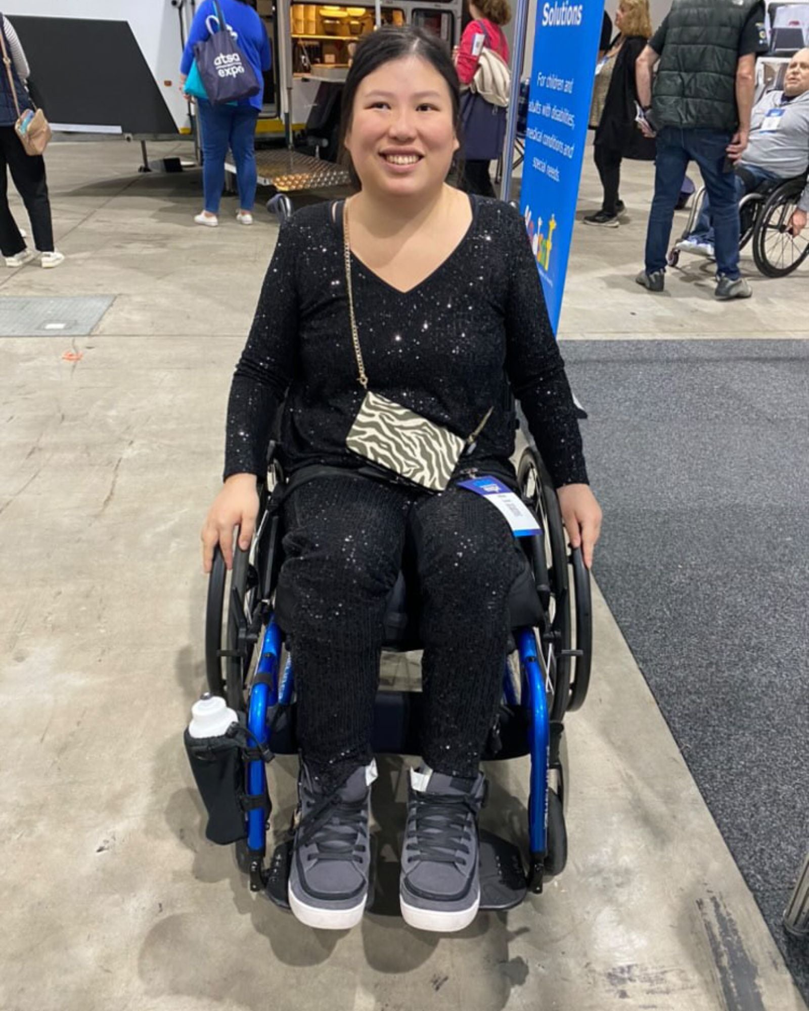 Suzie Q Seated Pants Sequins shown on a wheelchair user at an event, featuring a sparkly black sequined fabric. The adaptive pants are paired with a zebra-print crossbody bag and grey sneakers, demonstrating the stylish yet functional design perfect for wheelchair users.