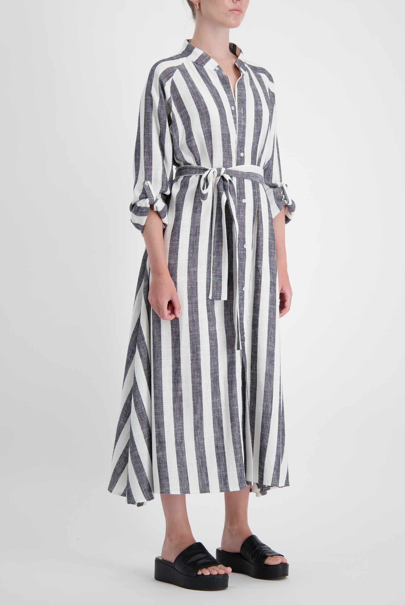 Sapphire Magnetic Shirt Dress in bold grey and white vertical stripes, featuring three-quarter rolled sleeves, collared neckline, and a waist-defining belt tie. The linen dress flows elegantly to midi length, shown paired with black platform sandals against a light background.