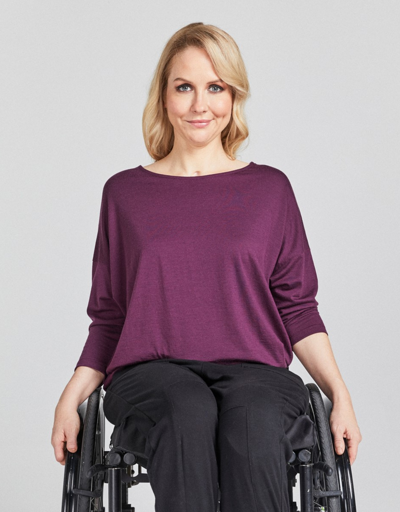 Blonde woman in a wheelchair wearing a grape (purple) round neck, loose top with 3/4 length sleeves and black pants. Forward facing. 