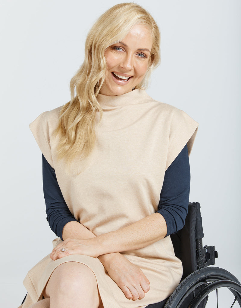 Image of a seated female in a wheelchair, with blonde hair wearing a natural cotton (beige colour) bat wing dress, with a high neckline. She has layered the top with a long sleeve navy blue shirt .Christina Stephens Adaptive Clothing Australia.