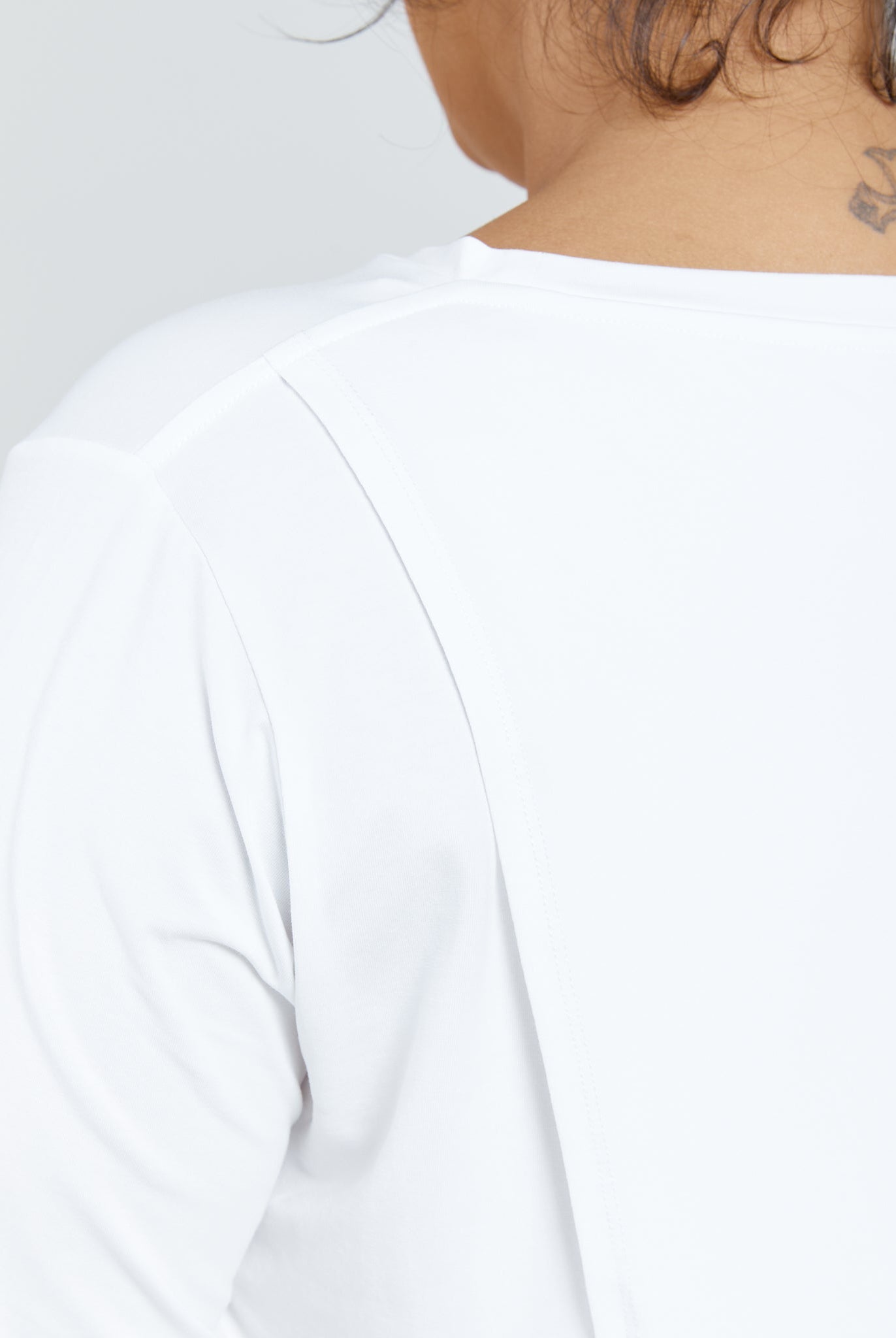 Elegant white Long Sleeve Leaf Back T-Shirt shown from behind, featuring a clean, minimalist design with a smooth shoulder seam detail. Small tattoo visible at neckline. Perfect casual-chic wardrobe essential photographed against a light background.