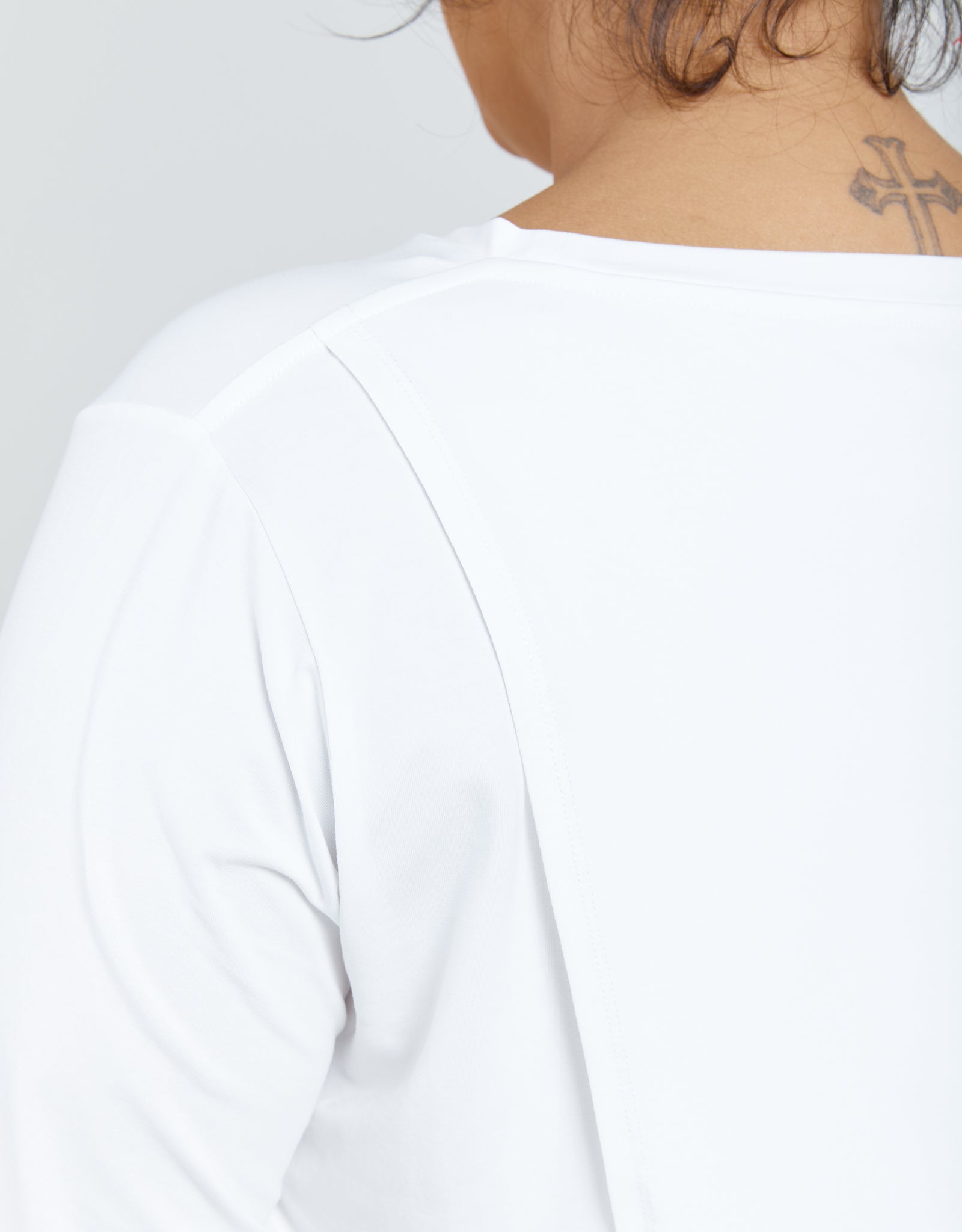 Elegant white Long Sleeve Leaf Back T-Shirt shown from behind, featuring a clean, minimalist design with a smooth shoulder seam detail. Small tattoo visible at neckline. Perfect casual-chic wardrobe essential photographed against a light background.