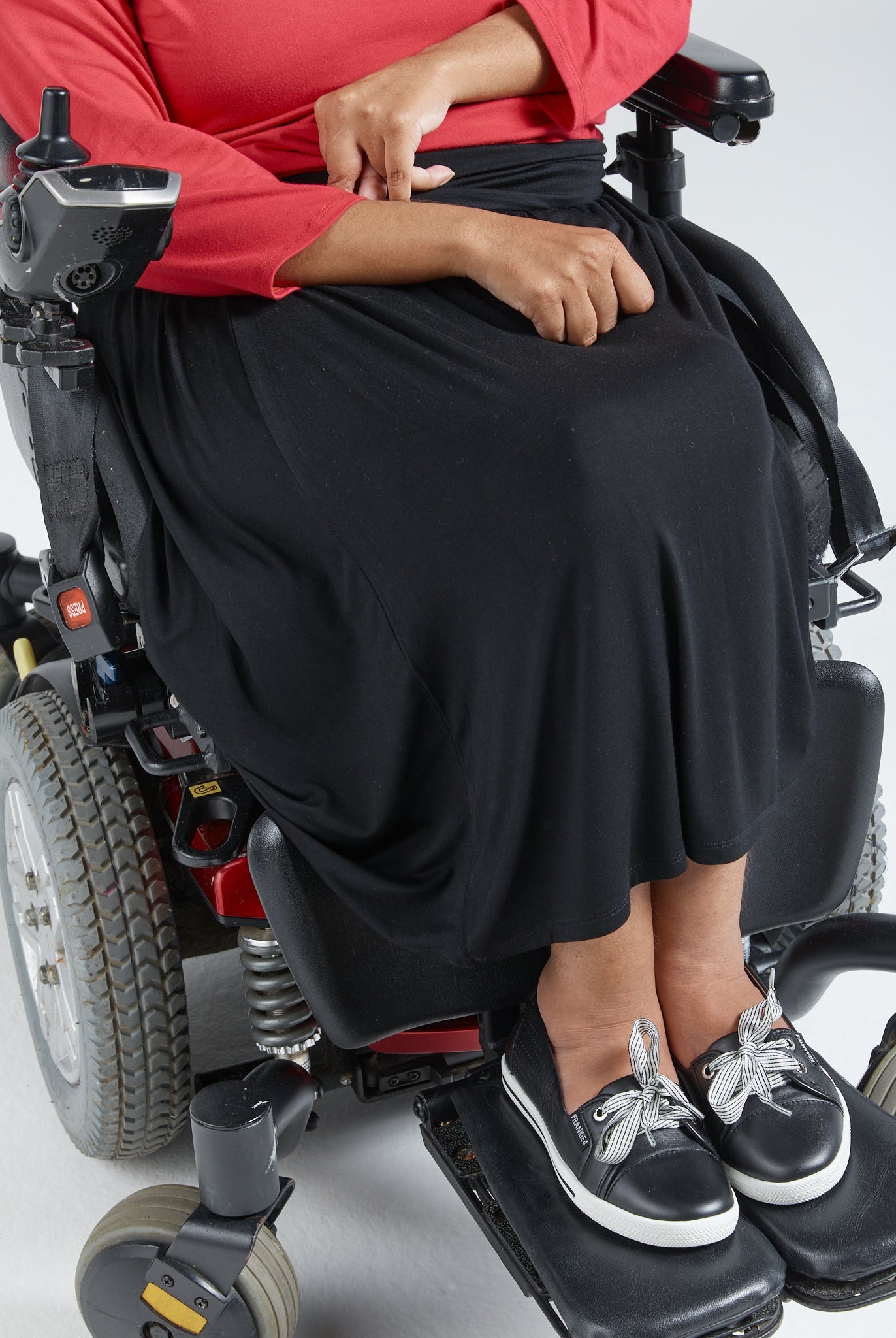 A-Line Skirt shown on a wheelchair user wearing a red top, demonstrating the skirt's easy-wear design with side pull tabs. The black stretchy skirt flows elegantly over the wheelchair seat, paired with casual black and white shoes.