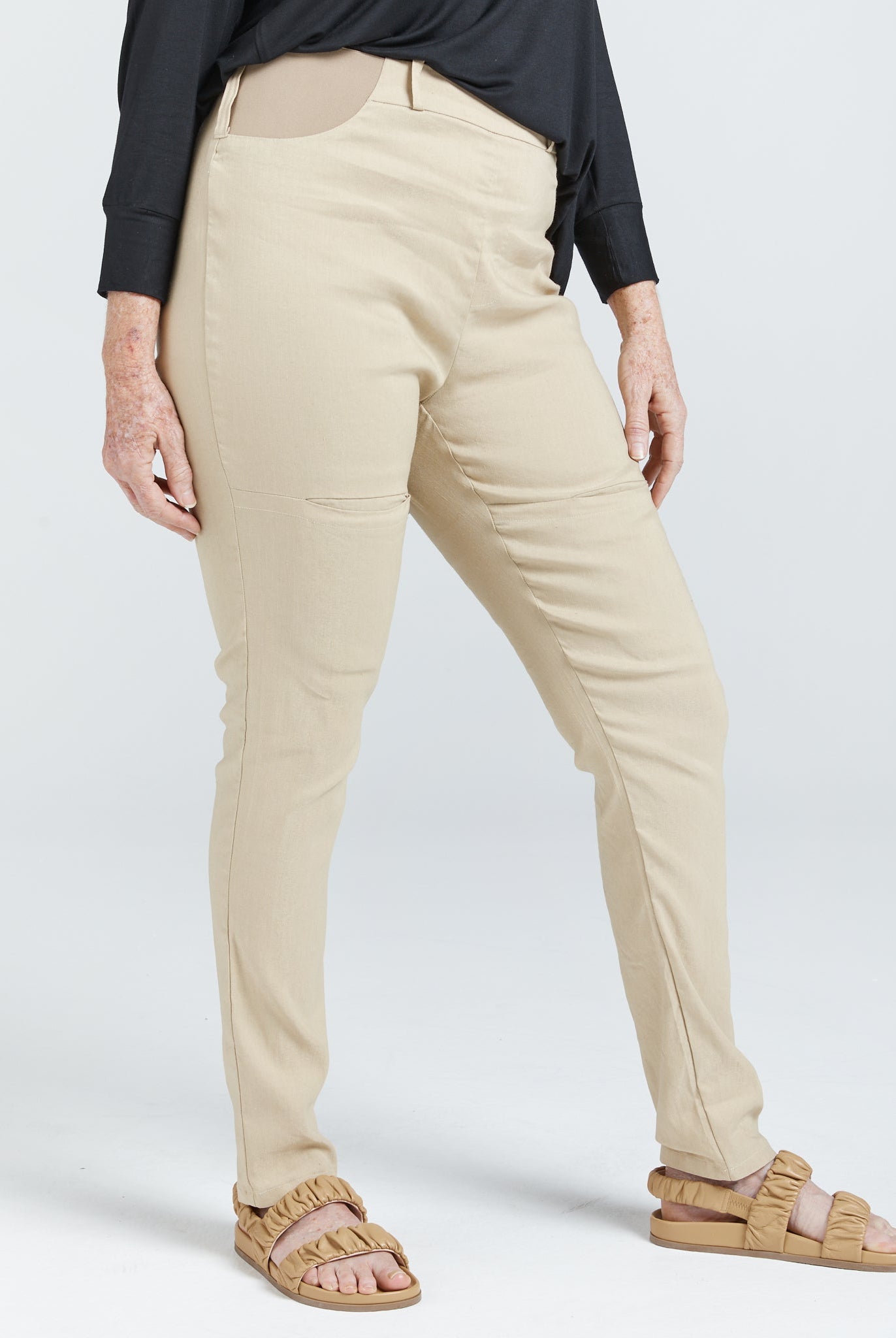 Seated Stretch Pant shown in beige color, paired with black top and tan sandals, featuring adaptive design with higher back rise and well-fitted leg length. Side view displays smooth fit and tailored silhouette against light background.