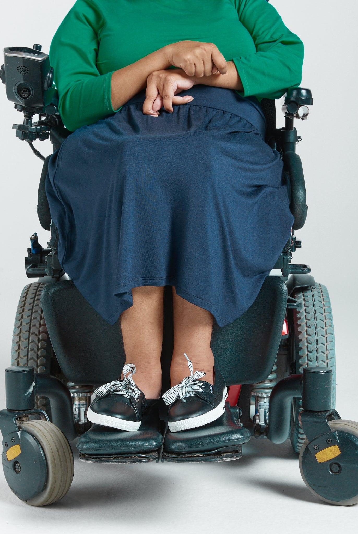 A-Line Skirt shown on a person seated in a power wheelchair, wearing a bright green top. The navy blue skirt drapes elegantly over the knees, demonstrating its adaptive design with a comfortable, stretchy fit. The image focuses on the accessible fashion features and practical styling.