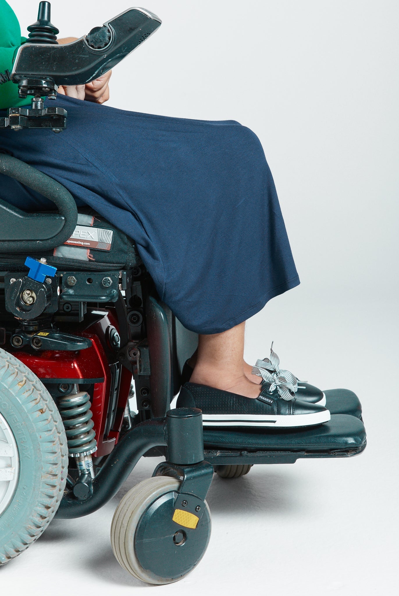A-Line Skirt shown on a power wheelchair user, featuring a flowing navy fabric draped over the wheelchair footrest. The adaptive skirt demonstrates easy-wear functionality, paired with casual footwear in a clinical setting against a white background.