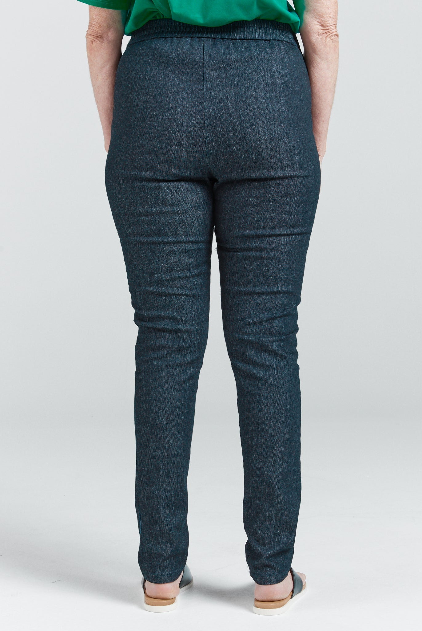 Seated Stretch Pant shown from back view, featuring dark denim fabric with elastic waistband and sleek tailored fit. Model wearing emerald green top, demonstrating the pants' full coverage design and smooth back panel construction.