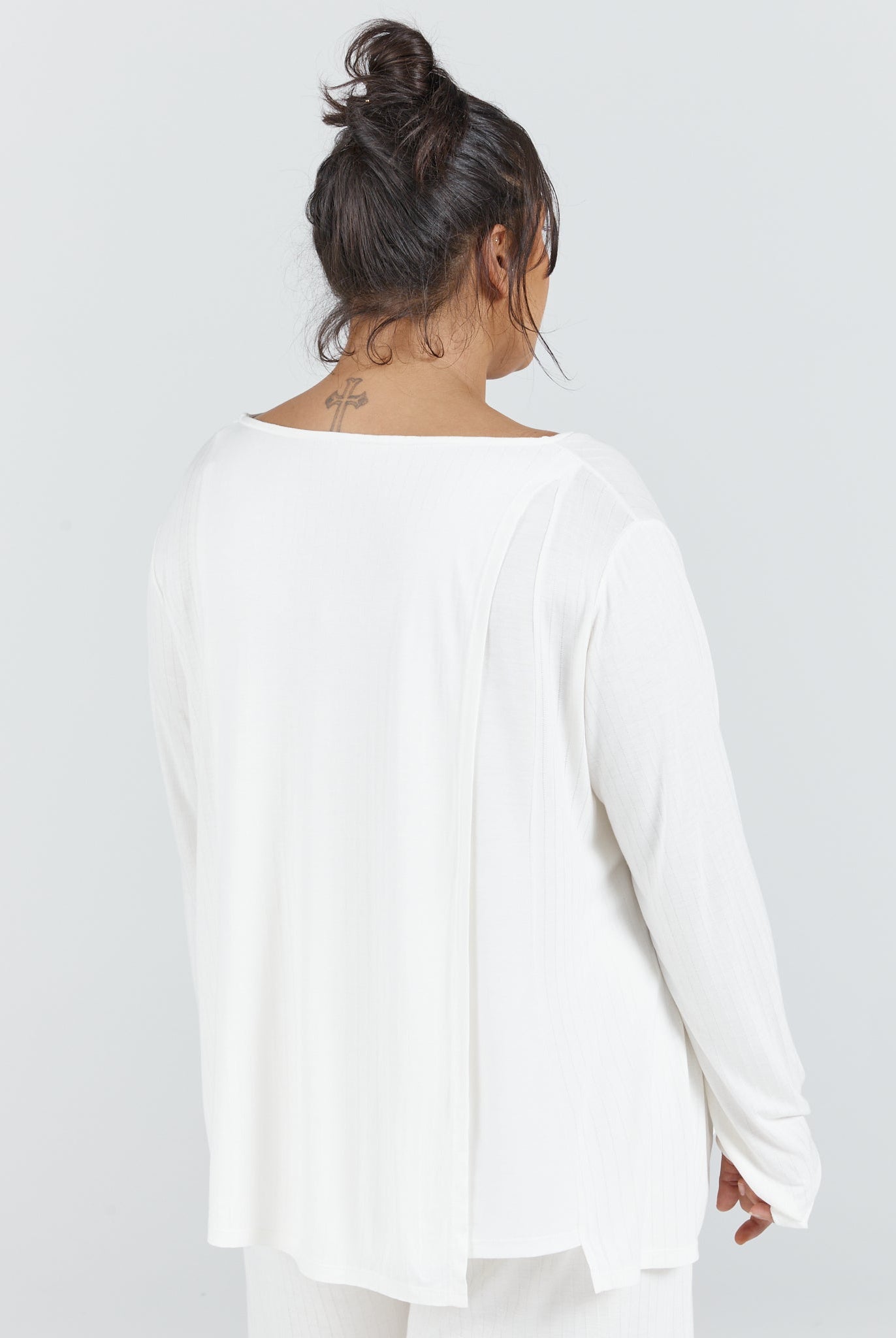 Essensual PJ Top shown from back view featuring a relaxed white long-sleeve design with boat neckline, loose-fitting silhouette, and soft draping fabric. Model wearing casual messy bun hairstyle against light gray background.