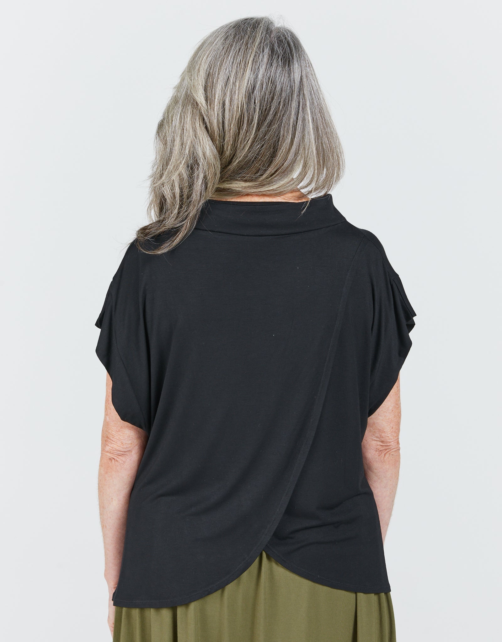 Leaf Back Blouse shown from behind, featuring elegant cross-over design in classic black with short flutter sleeves. The loose-fitting garment drapes gracefully, highlighting the Asian-inspired back detail against a light background. Gray hair showcases the neckline and shoulder construction.