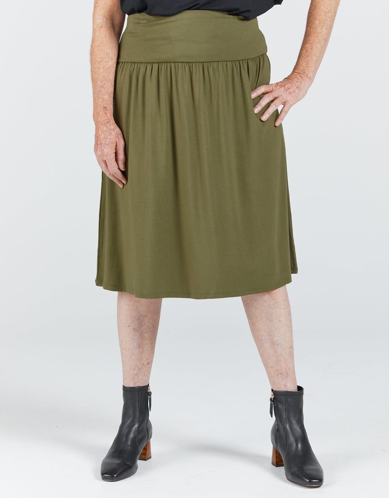 Standing lady wearing a khaki bamboo skirt that reaches past her knees. It has a wide waist band and gathers below the band. She is wearing a black top and boots. Christina Stephens Adaptive Clothing Australia. 