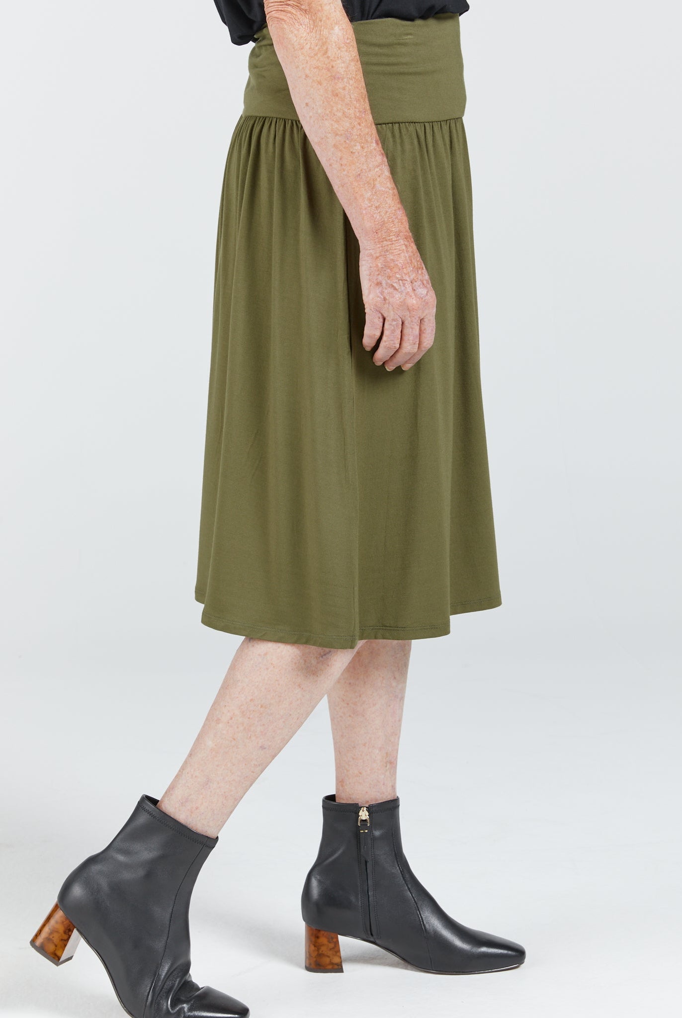 A-Line Skirt in olive green bamboo fabric shown from side view, featuring a comfortable wide waistband and flowing midi-length silhouette paired with black ankle boots. Skirt demonstrates stretchy, pull-on design without visible fastenings.