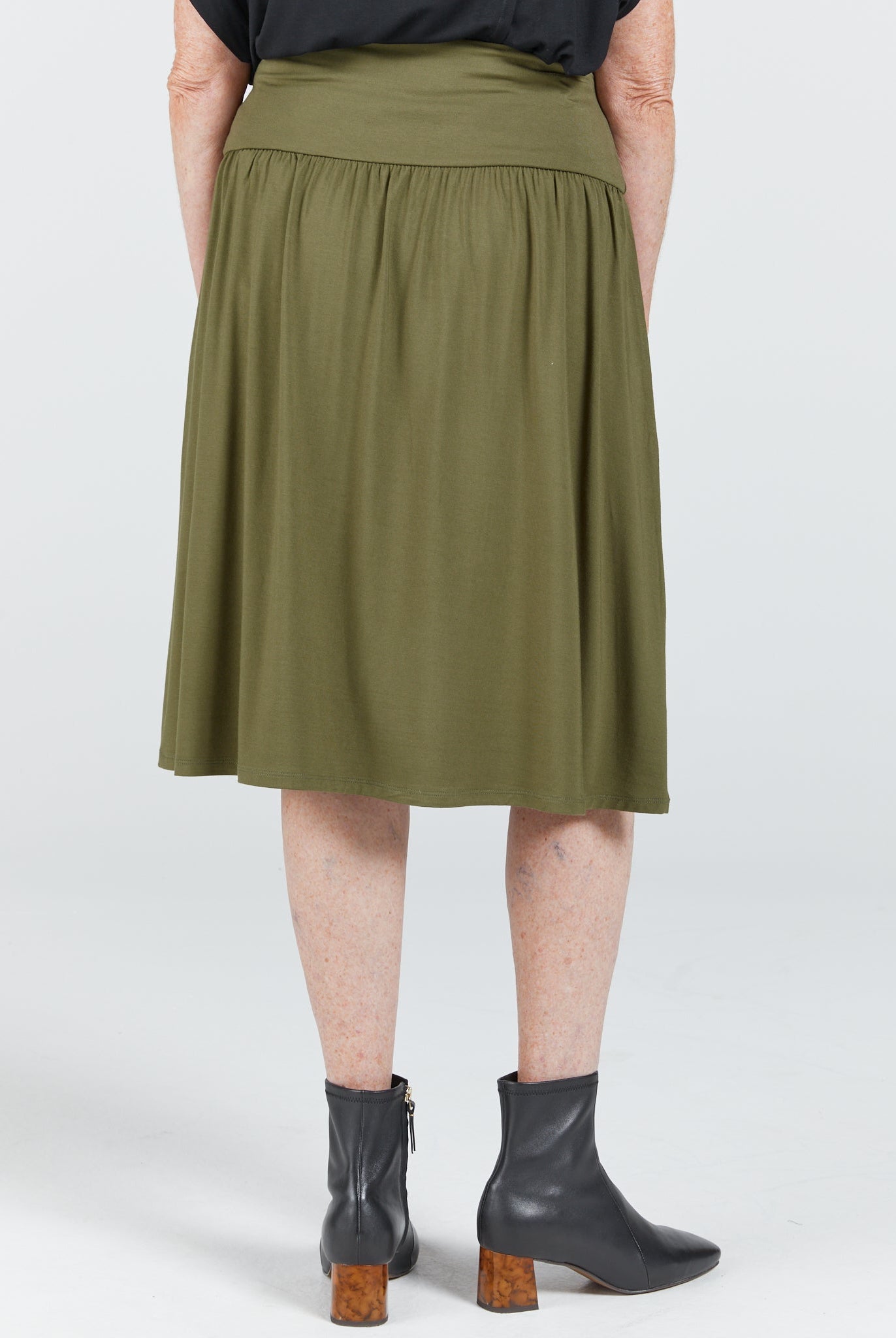 Olive green A-Line skirt shown from back view, featuring a wide waistband and flowing bamboo fabric that falls just below the knee. Side pull tabs visible at waist for easy dressing. Paired with black boots and dark top.