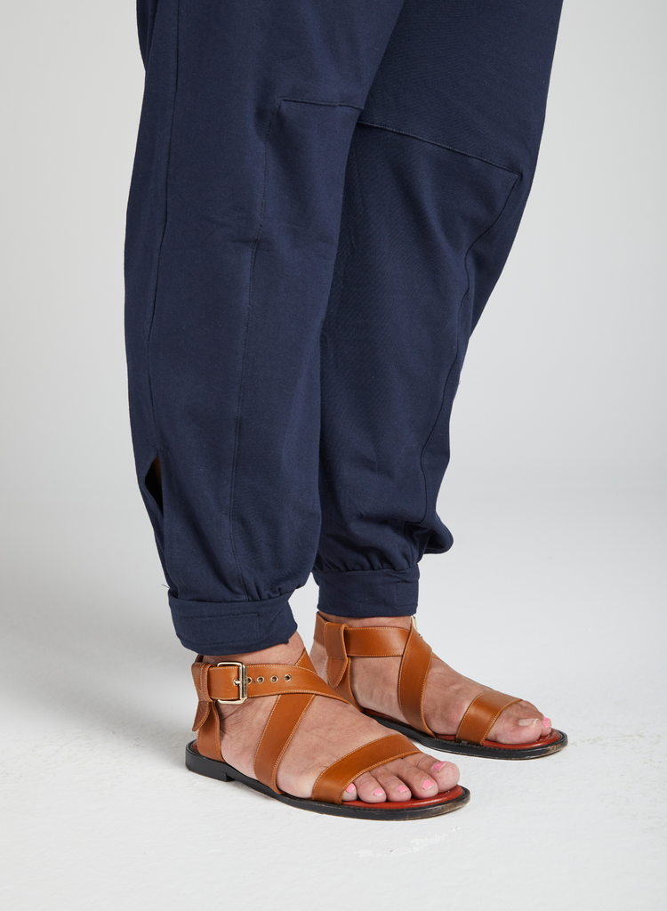 Model is wearing navy pants and tan sandals. Pants feature rectangular seams on the inner side of both legs. Additional cuffs on both leg openings.