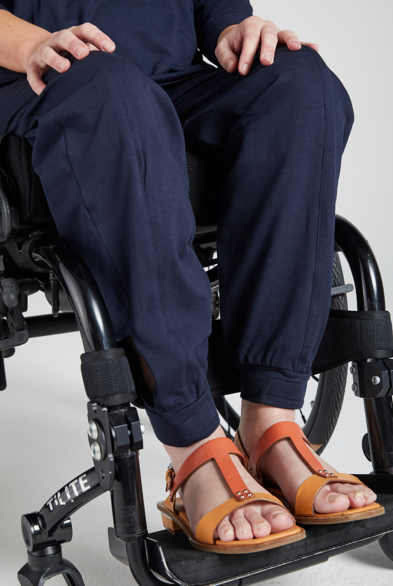 Tapered Track Pants with Hidden Pouch shown on a seated individual in a wheelchair, displaying navy blue pants with tapered legs. The image focuses on the lower body, highlighting the pants' fit and style, paired with tan leather sandals.