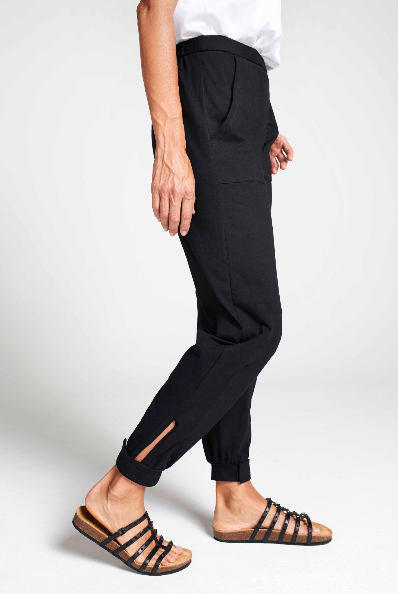 Tapered Track Pants with Hidden Pouch shown in black, featuring side view of slim-fit design with ankle slit detail, paired with white top and strappy sandals against light gray background, highlighting comfortable tapered silhouette and practical hidden pouches.