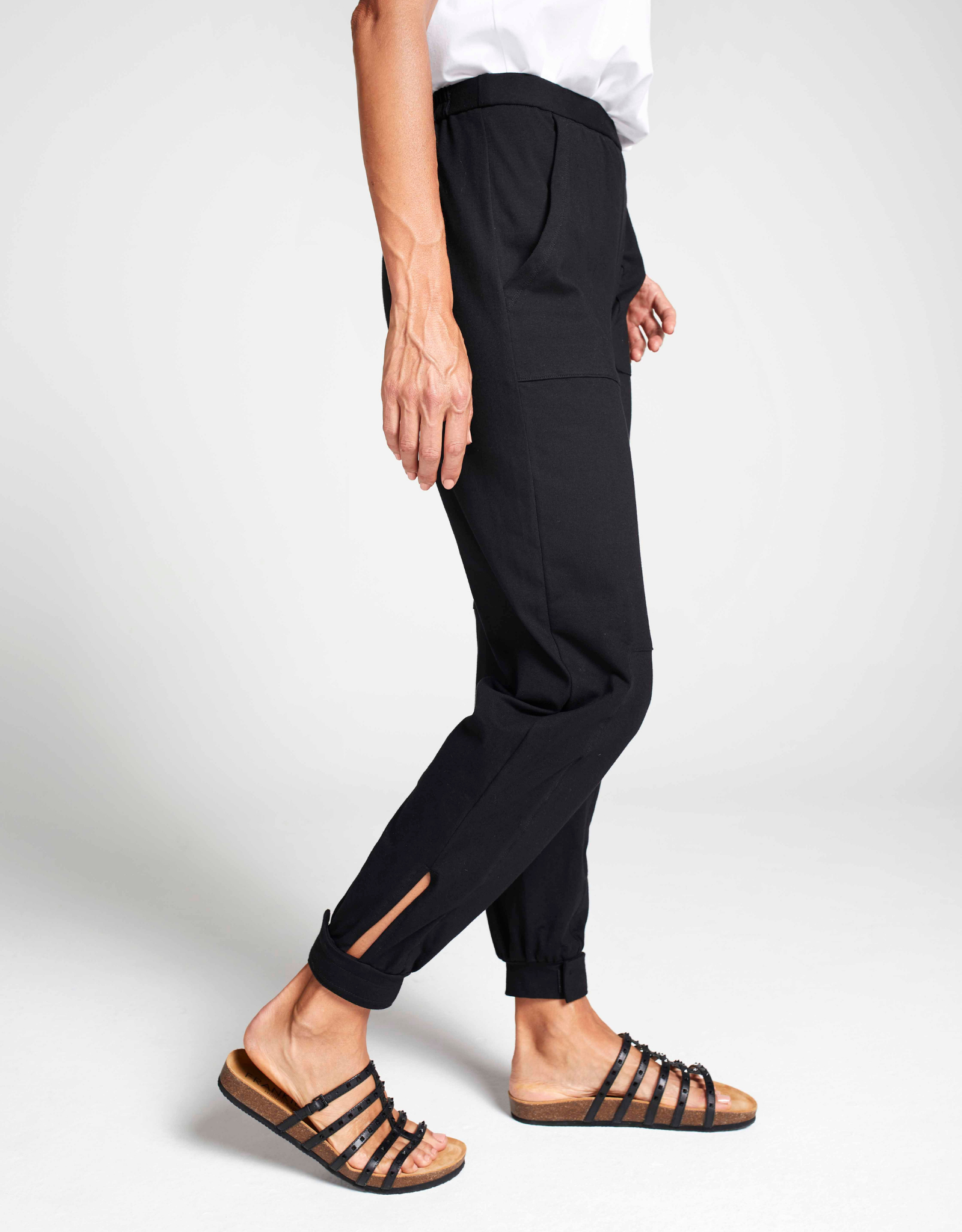 Tapered Track Pants with Hidden Pouch shown in black, featuring side view of slim-fit design with ankle slit detail, paired with white top and strappy sandals against light gray background, highlighting comfortable tapered silhouette and practical hidden pouches.