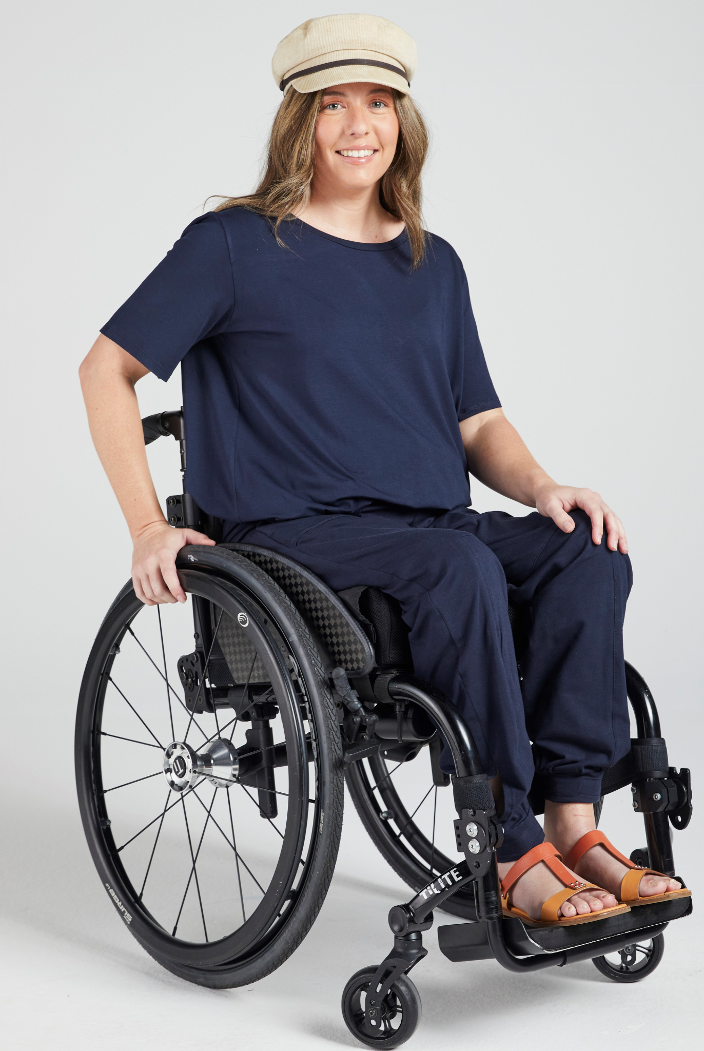 Model wearing Short Sleeve Leaf Back T-Shirt in navy blue, styled with beige cap and orange sandals, seated in a modern wheelchair against light grey background, demonstrating adaptive fashion and inclusive design.