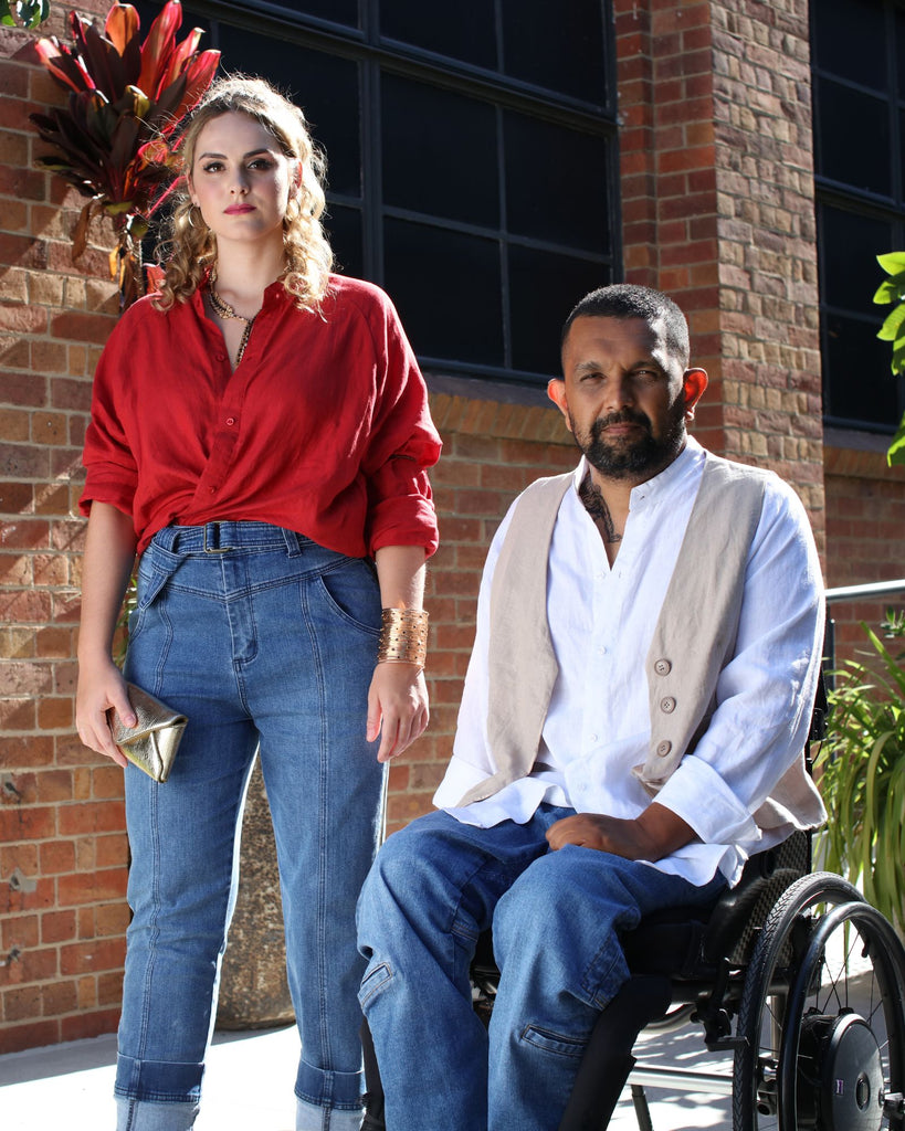 A female standing with her back to a brick wall wearing a red linen shirt tucked into blue denim jeans with zippers up both side seams. She is standing next to a seated male in a wheelchair. He has dark skin and dark hair, and is wearing a beige linen vest, white linen shirt and blue jeans. Christina Stephens Adaptive Clothing Australia. 