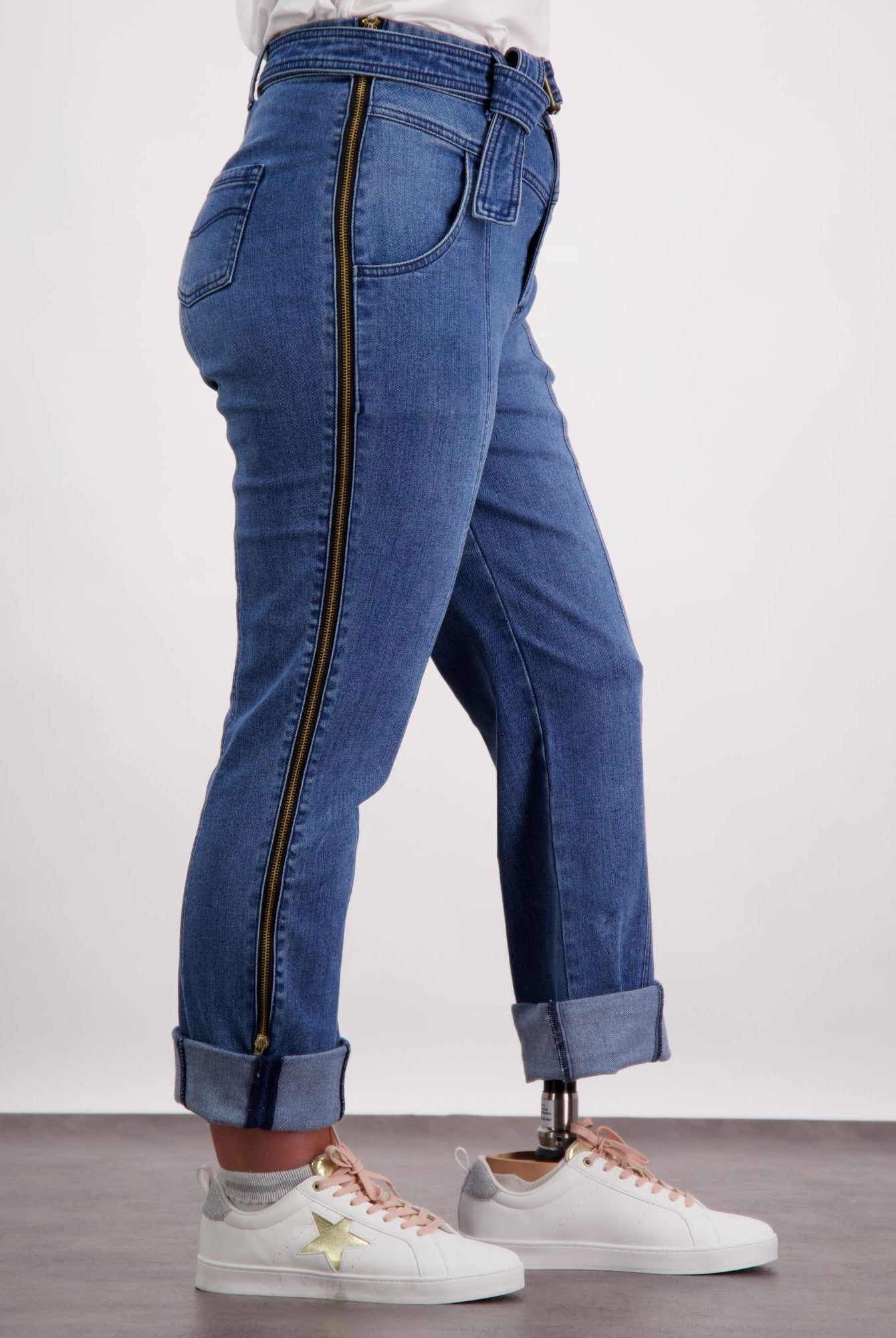 Christina Prosthetic Jeans featuring full-length side zippers, shown in medium wash denim with cuffed ankles. Adaptive design showcases innovative tear-resistant fabric and accessibility features, paired with white sneakers and a casual white top.