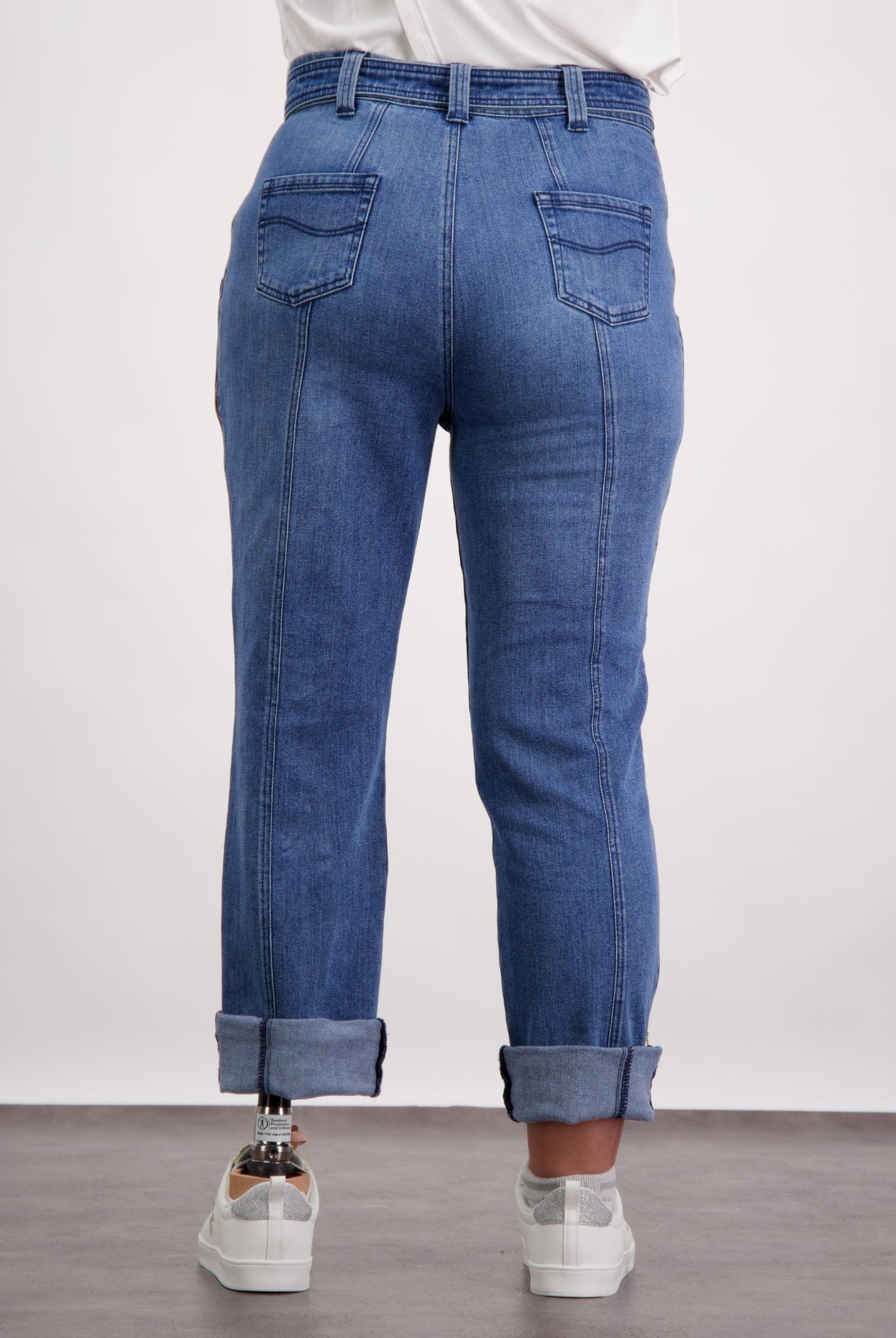 Christina Prosthetic Jeans shown from back view, featuring medium-wash denim with reinforced stitching, straight-leg design, and side zippers. Jeans are rolled at ankles, styled with white sneakers and casual white top. Demonstrates adaptive fashion functionality.