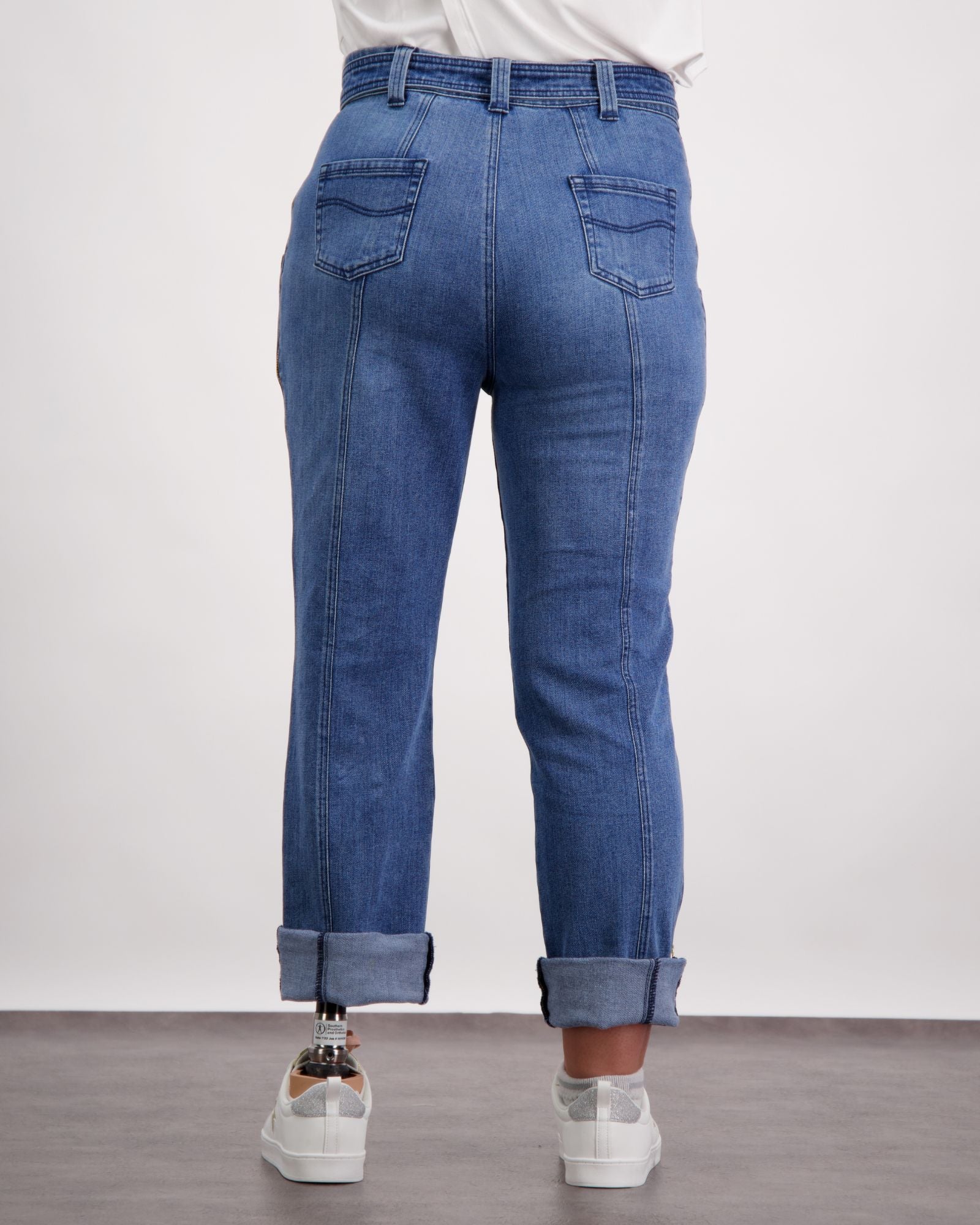 Christina Prosthetic Jeans shown from back view, featuring medium-wash denim with reinforced stitching, straight-leg design, and side zippers. Jeans are rolled at ankles, styled with white sneakers and casual white top. Demonstrates adaptive fashion functionality.