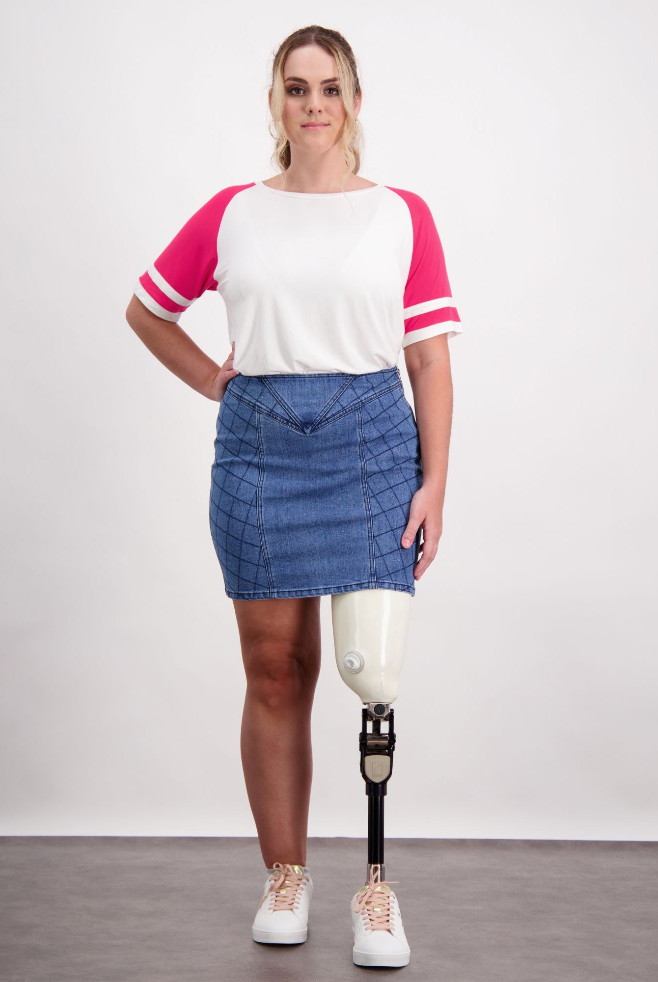 Bella Skirt shown in quilted blue denim with unique stitching pattern, paired with white and pink baseball-style top. Model wearing casual sneakers demonstrates the skirt's adaptive design features including dual side zippers, standing against light background.