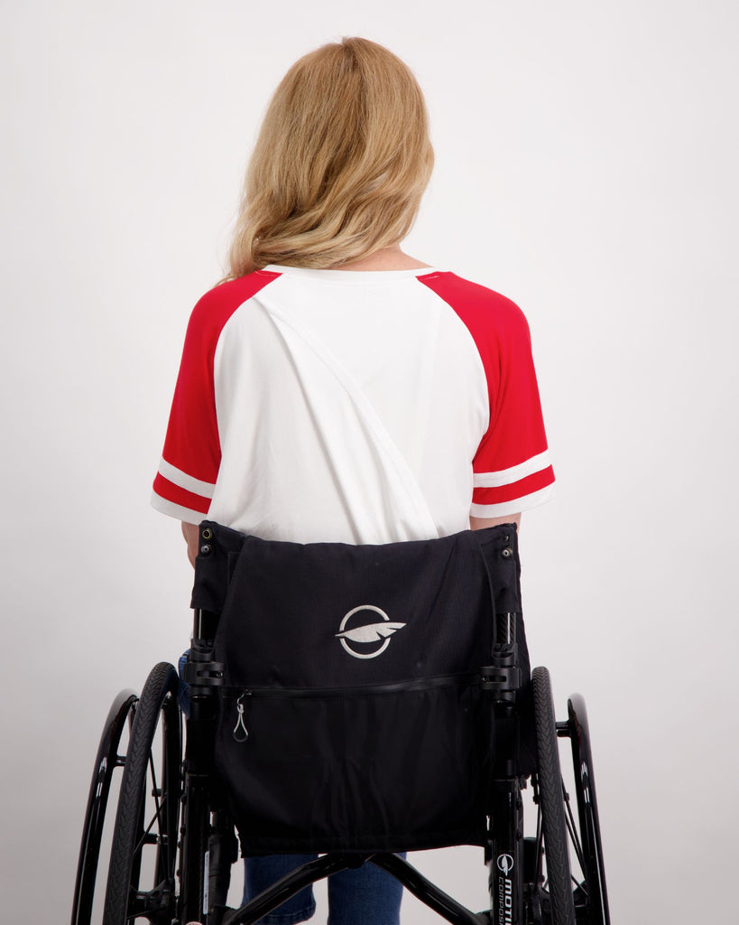 A blonde female seated in a wheelchair is wearing a white and red raglan t-shirt. She is facing backwards to show the leaf back split on the t-shirt. The leaf back split is slightly opened. She is sitting in a black coloured wheelchair. Christina Stephens Australian Adaptive Clothing.